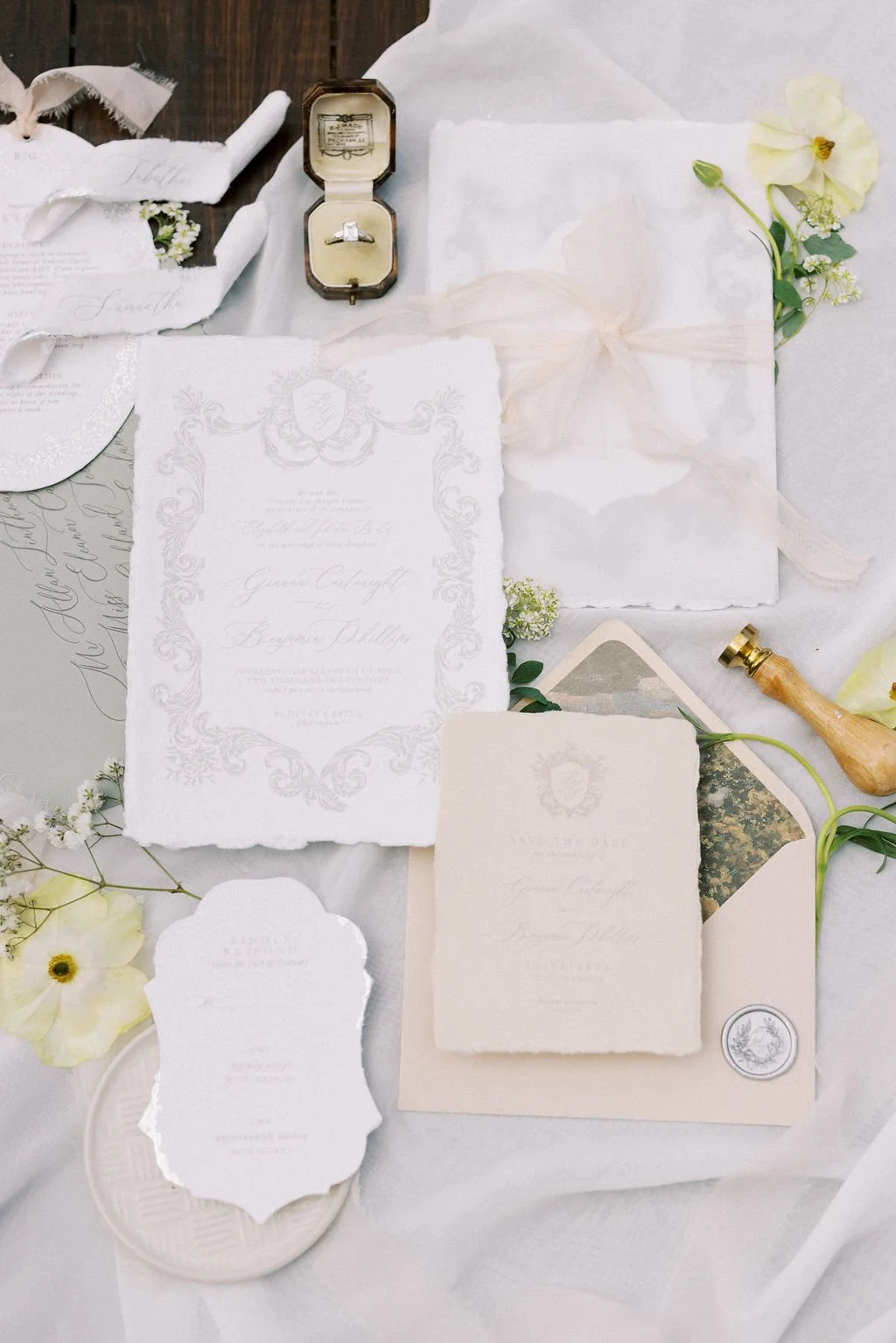 An assortment of elegant wedding invitation papers, a small ring box with a diamond engagement ring, a wax stamp, and decorative flowers arranged on a white fabric surface.
