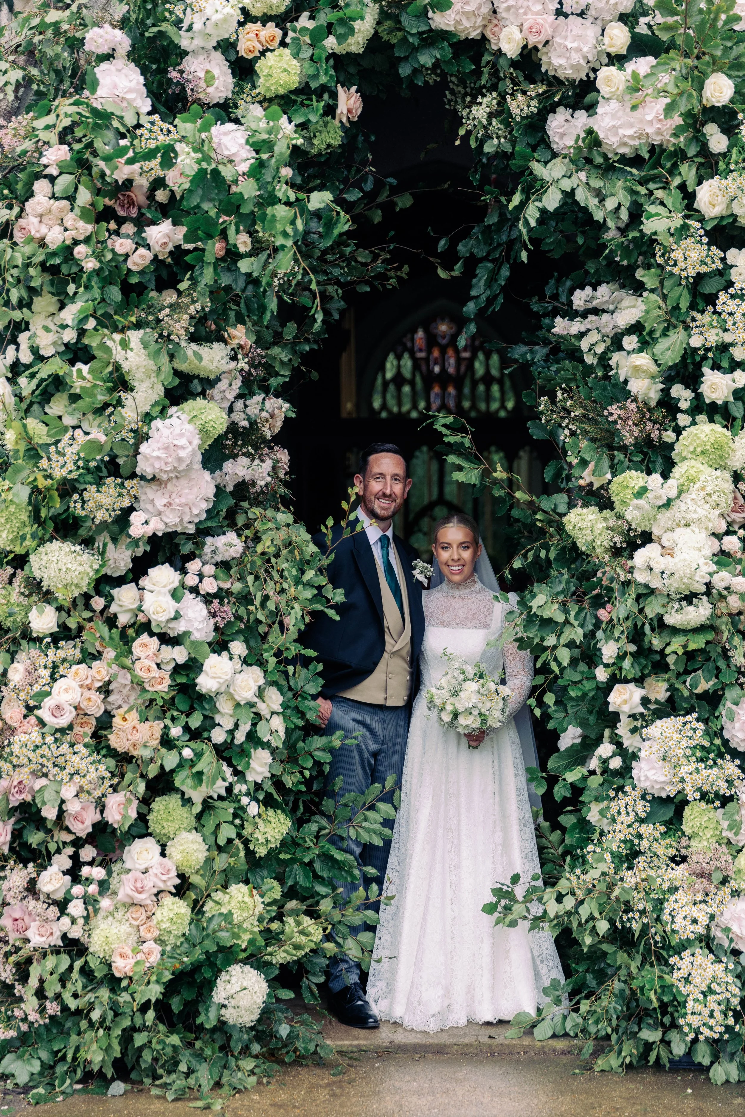 A bride and groom standing in front of a floral arch on their wedding day, inside a church. The arch is decorated with summer flowers. The bride is holding a bouquet and wearing a lace wedding dress, the groom is in a suit with a vest and tie, both s