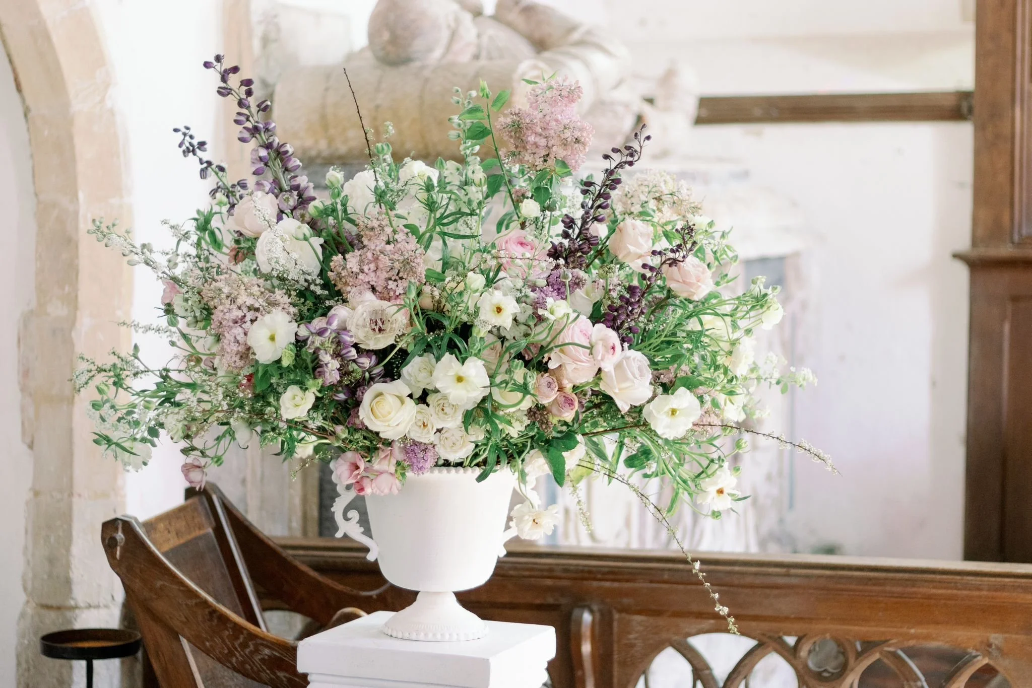 A large floral arrangement in a white urn-style vase containing pink, white, and purple flowers, with greenery, placed on a white pedestal in a room with a brick and plaster wall and wooden furniture.