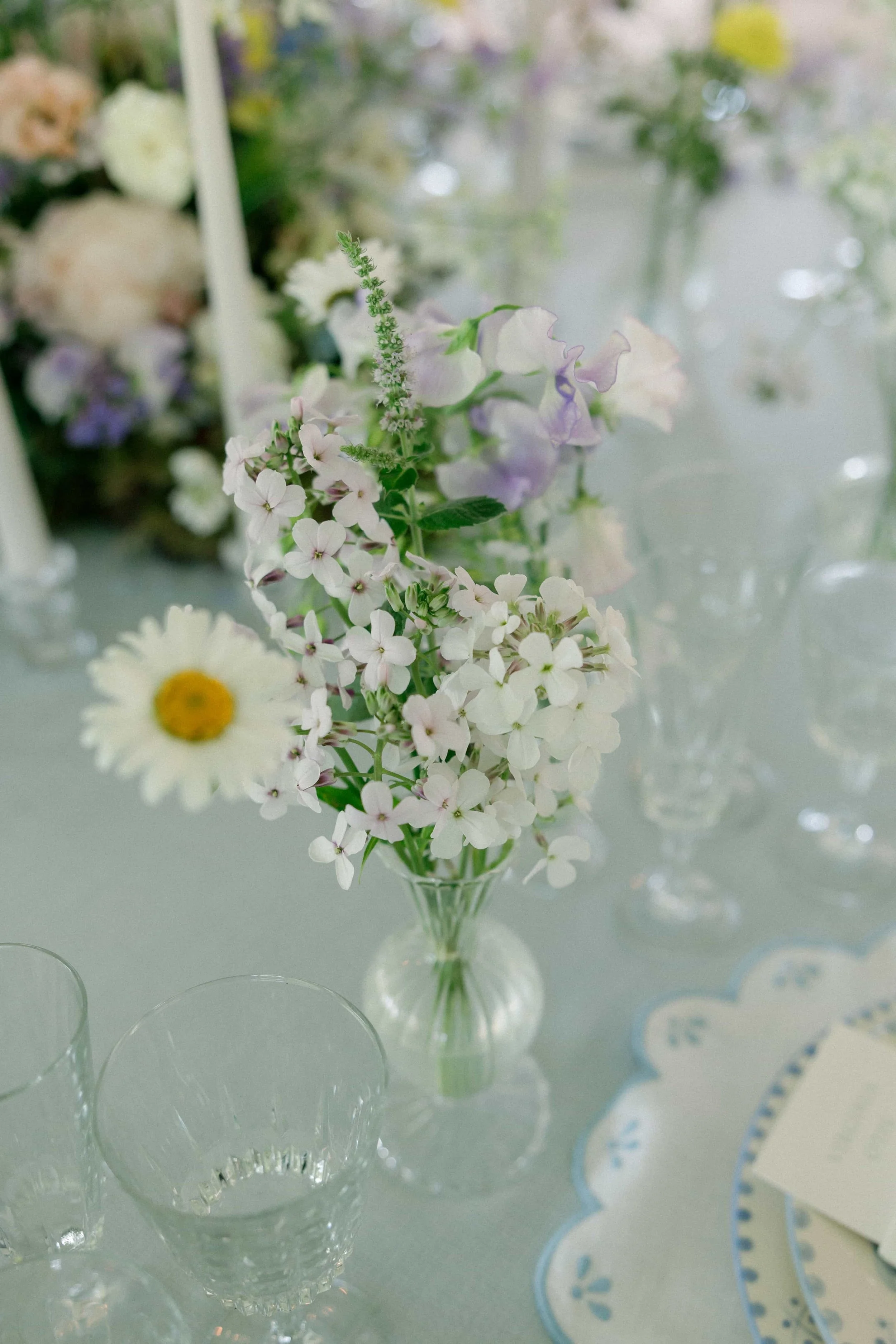 Close-up of a bud vase of white, purple, and light pink flowers in a glass vase, on a table, with clear glassware and a paper placemat visible. The Botany House Florist at Wilderness Reserve, Planned by Attabara