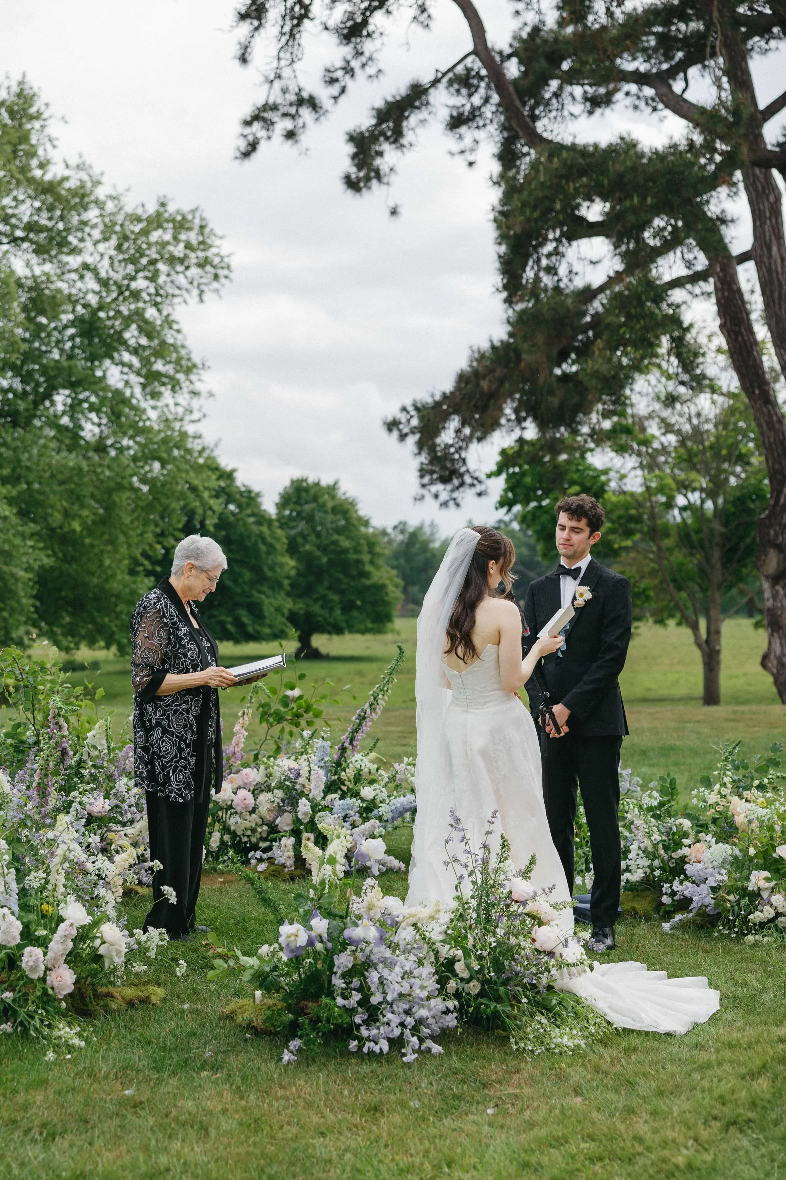 A wedding ceremony outdoors with a bride and groom standing in front of an officiant, surrounded by flowers and greenery. Wilderness Reserve, Suffolk Florals By The Botany House