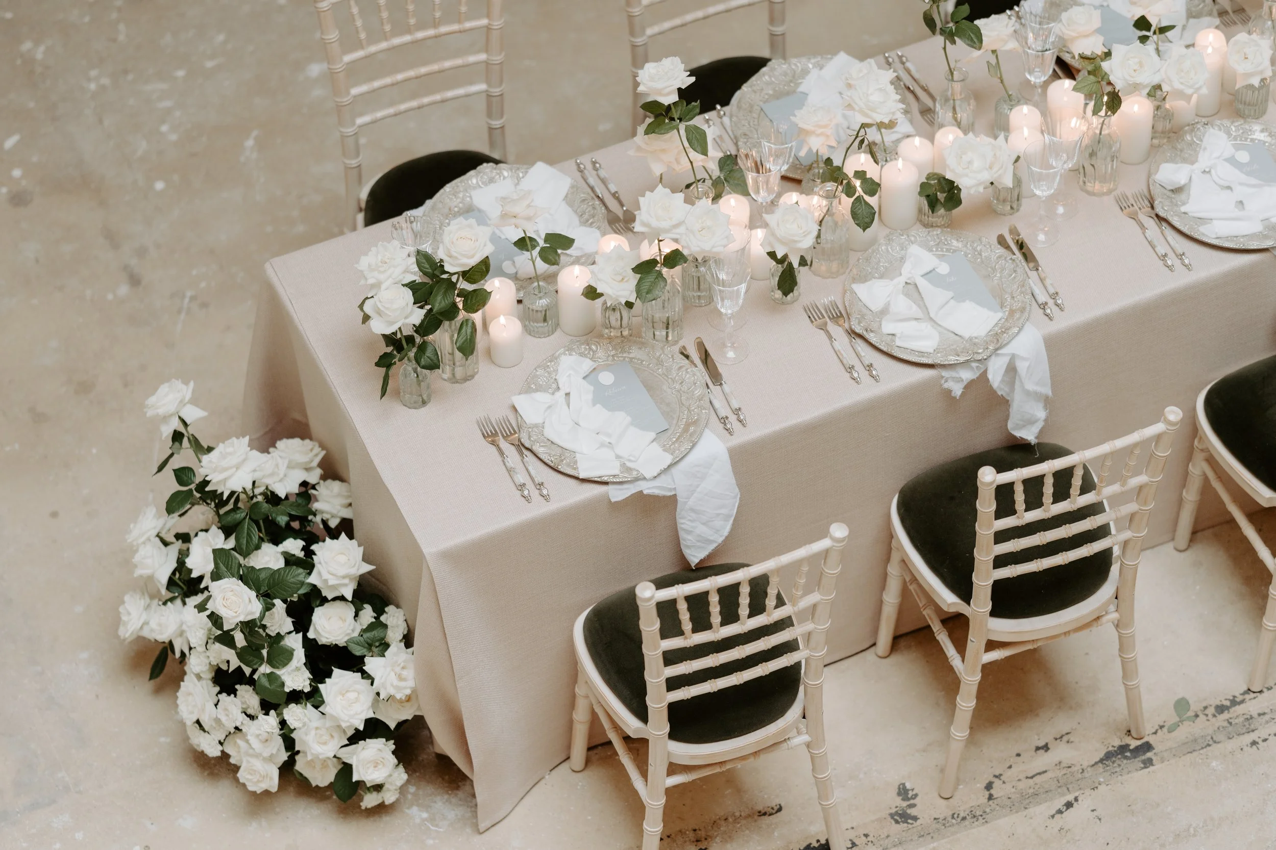 Elegant dining table decorated with white roses, candles, glassware, and silverware, set for a formal occasion with black cushioned chairs surrounding it.
