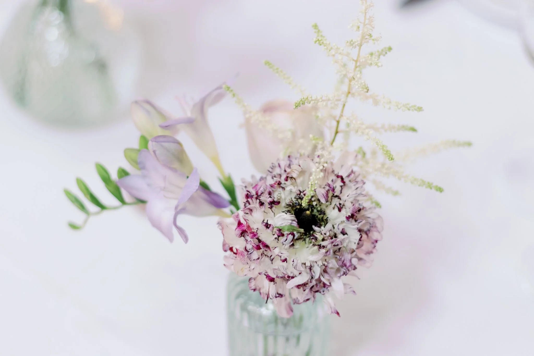 A delicate bouquet of light purple and white flowers in a glass vase on a soft white background.