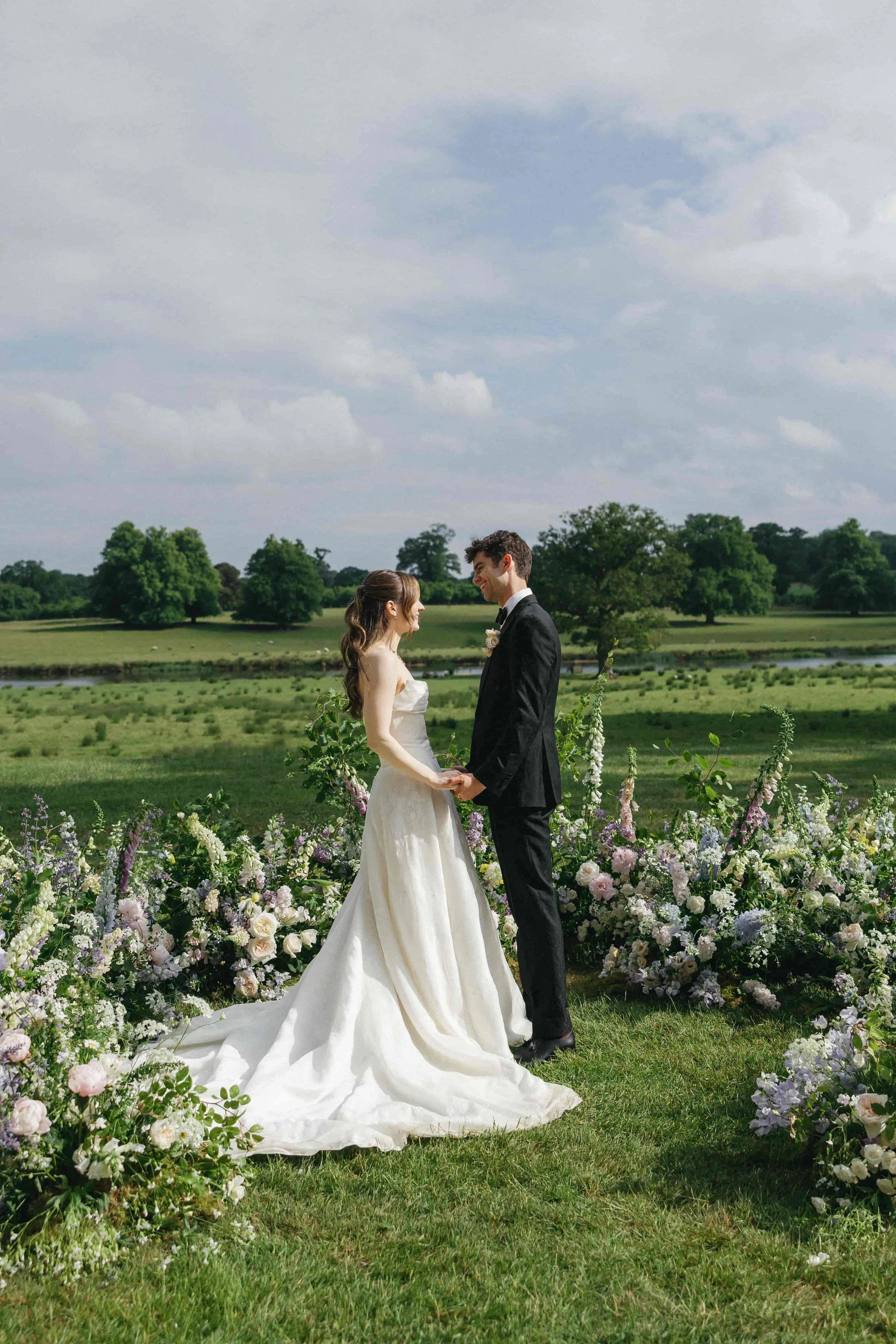 Bride and groom during outdoor ceremony surrounded by floral arrangements and guests watching in green meadow. The Botany House Wedding Florist. Planner Attabara Studio