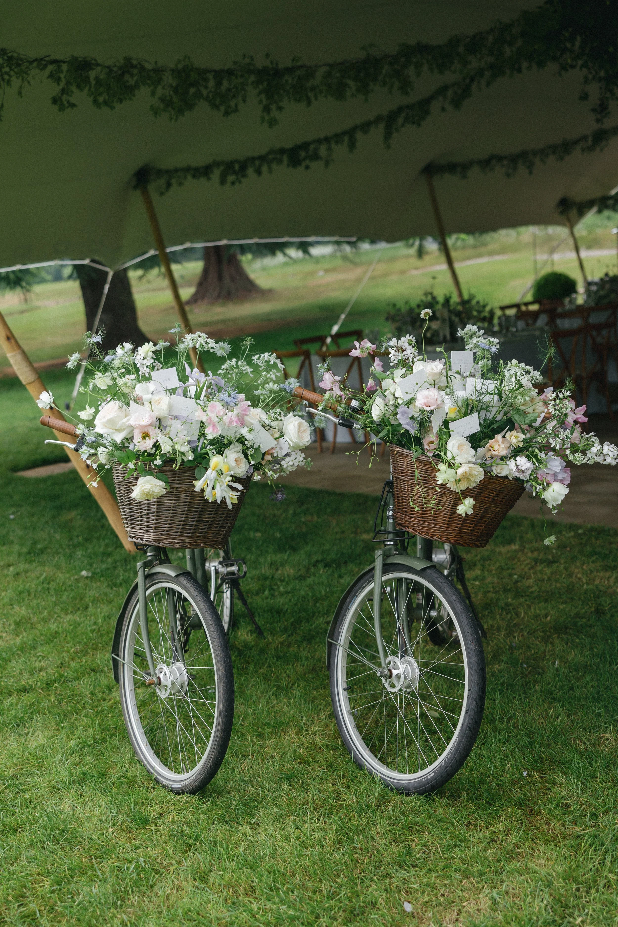 Two bicycles with baskets filled with white and pink flowers parked on grass under a large green canopy at an outdoor event.