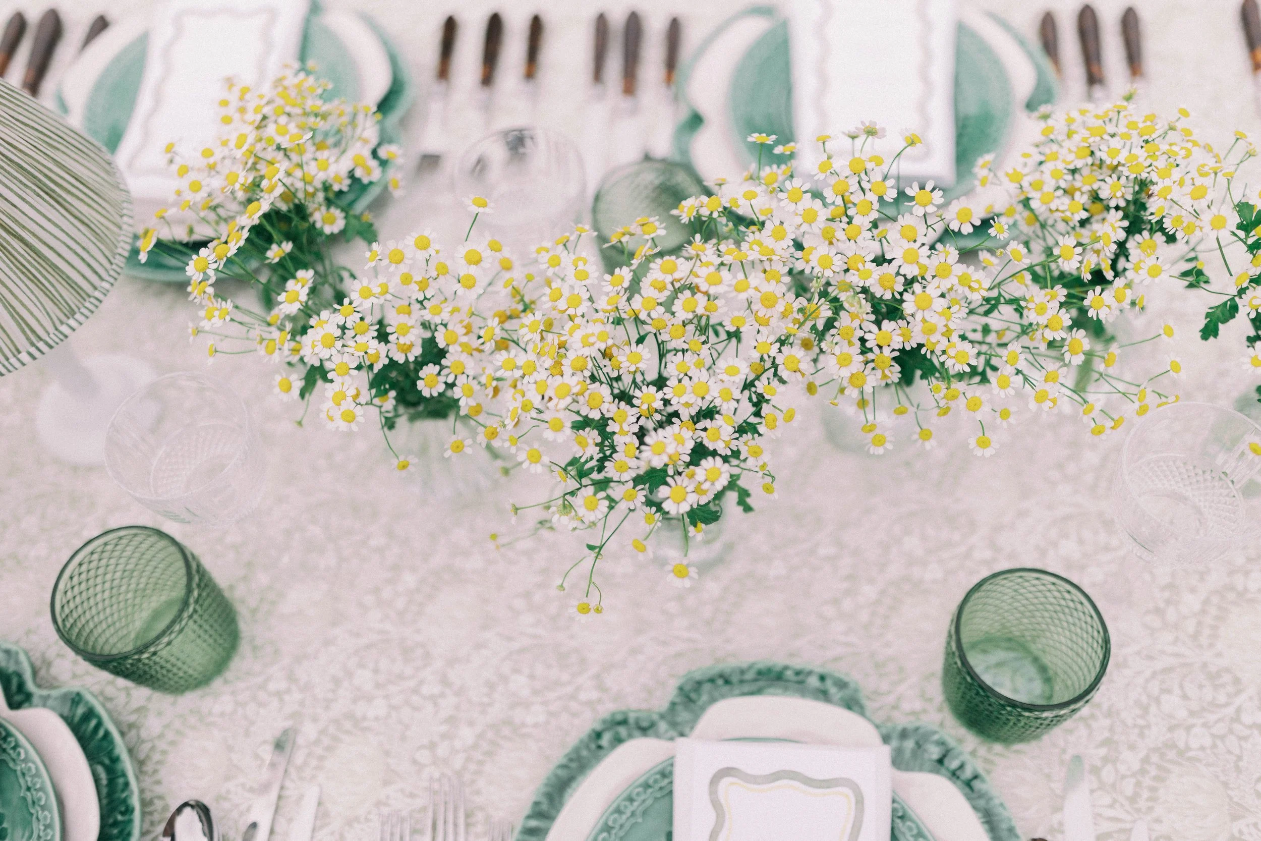 A table setting with a centerpiece of small white and yellow daisies, surrounded by green glassware and pale green and white dishware with napkins, set on a floral white tablecloth.