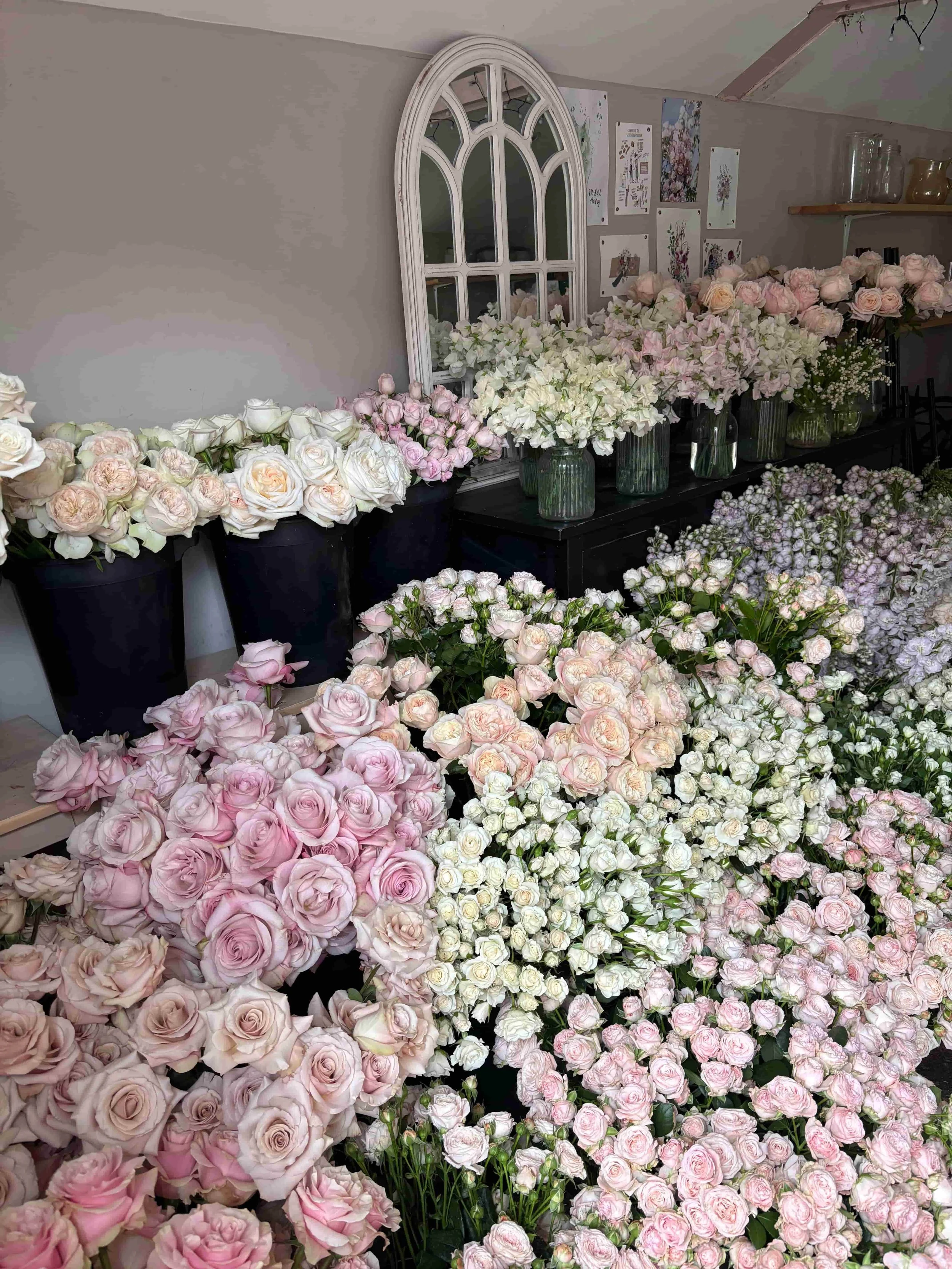 A room filled with various pink and white roses arranged in black and glass vases and piled on tables and the floor, floral artwork in the background. The Botany House studio in Norfolk. In preparation for Mother's Day bouquet gifting, the best in No