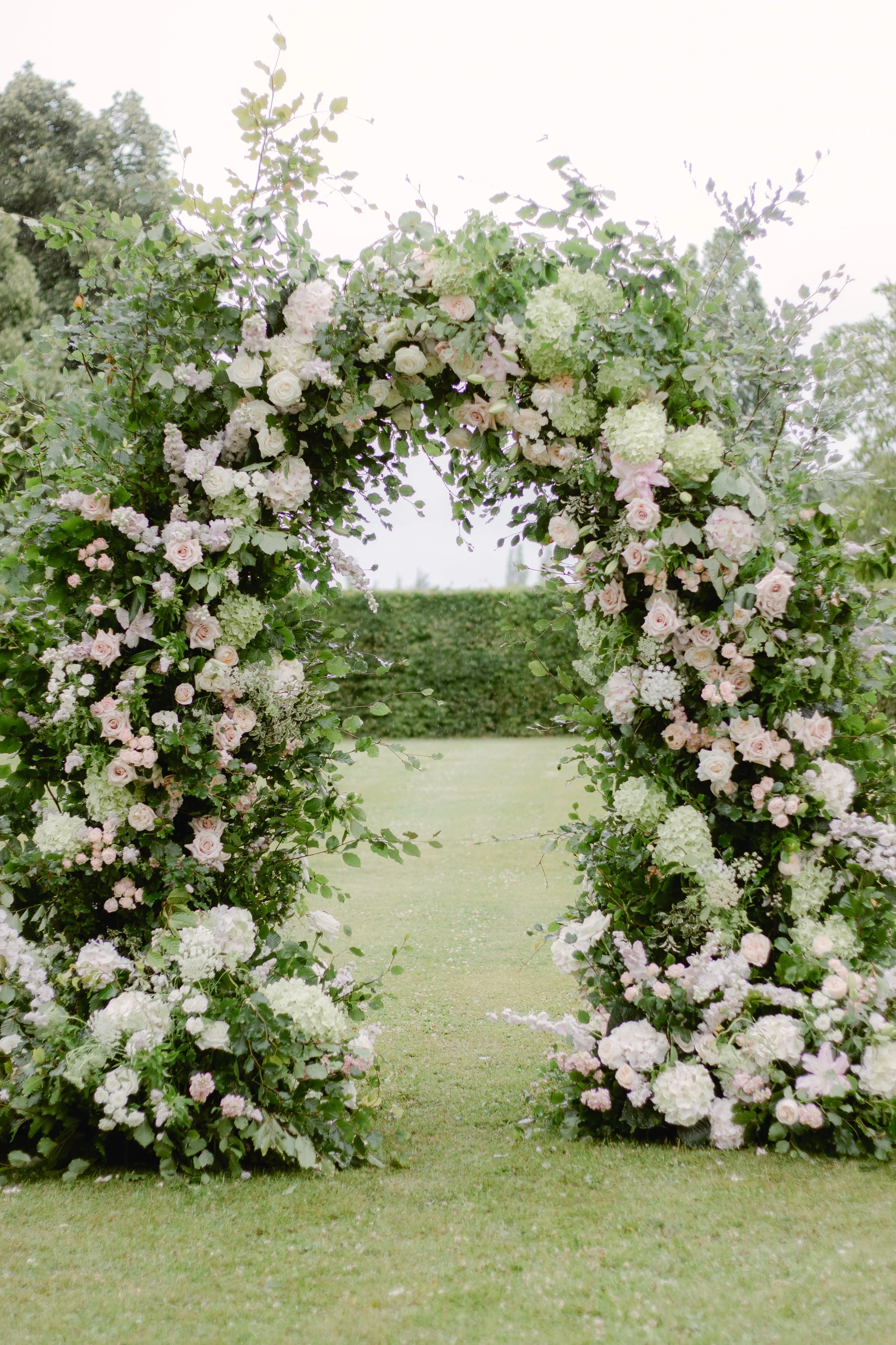 Flower arch decorated with pink and white roses, hydrangeas, and greenery, set outdoors on a grassy area.