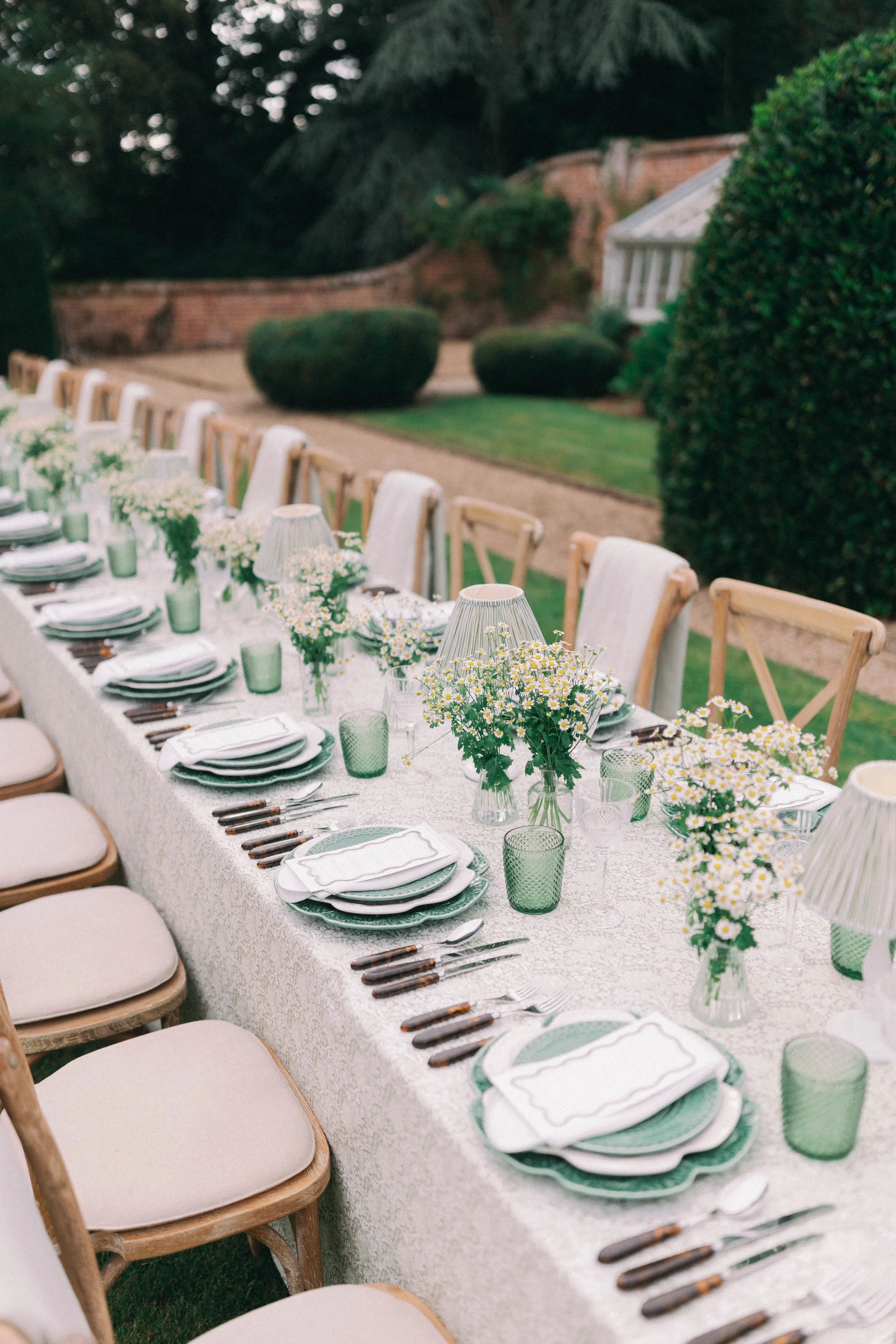 Long outdoor dining table decorated with white tablecloth, floral centerpieces, tableware, and lamps, set up in a countryside garden.