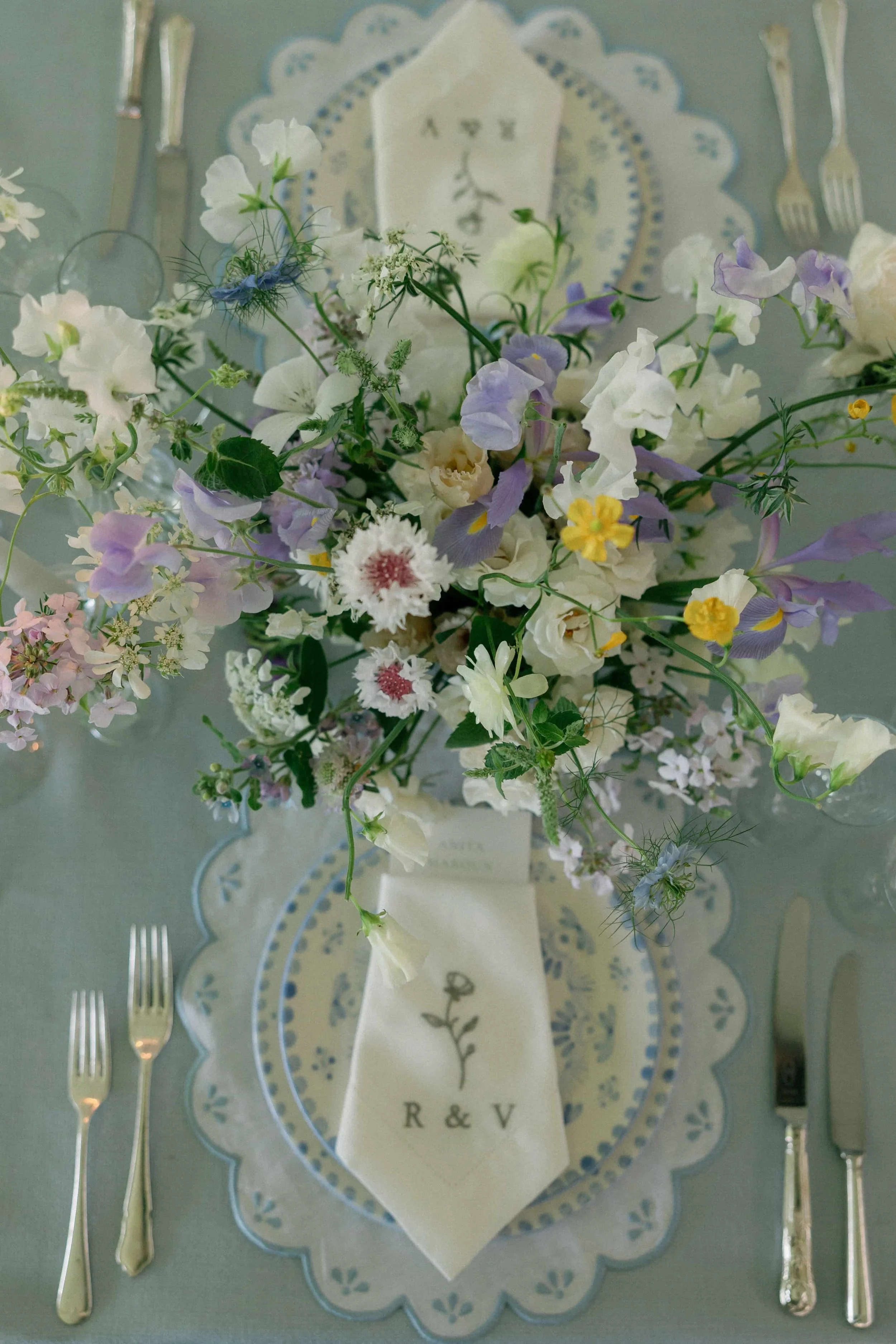 A table setting with a floral centerpiece, place settings, and cutlery. The flowers are white, purple, yellow, and pink, arranged on a scalloped placemat, with a cloth napkin that has an embroidered flower and the initials R & V.