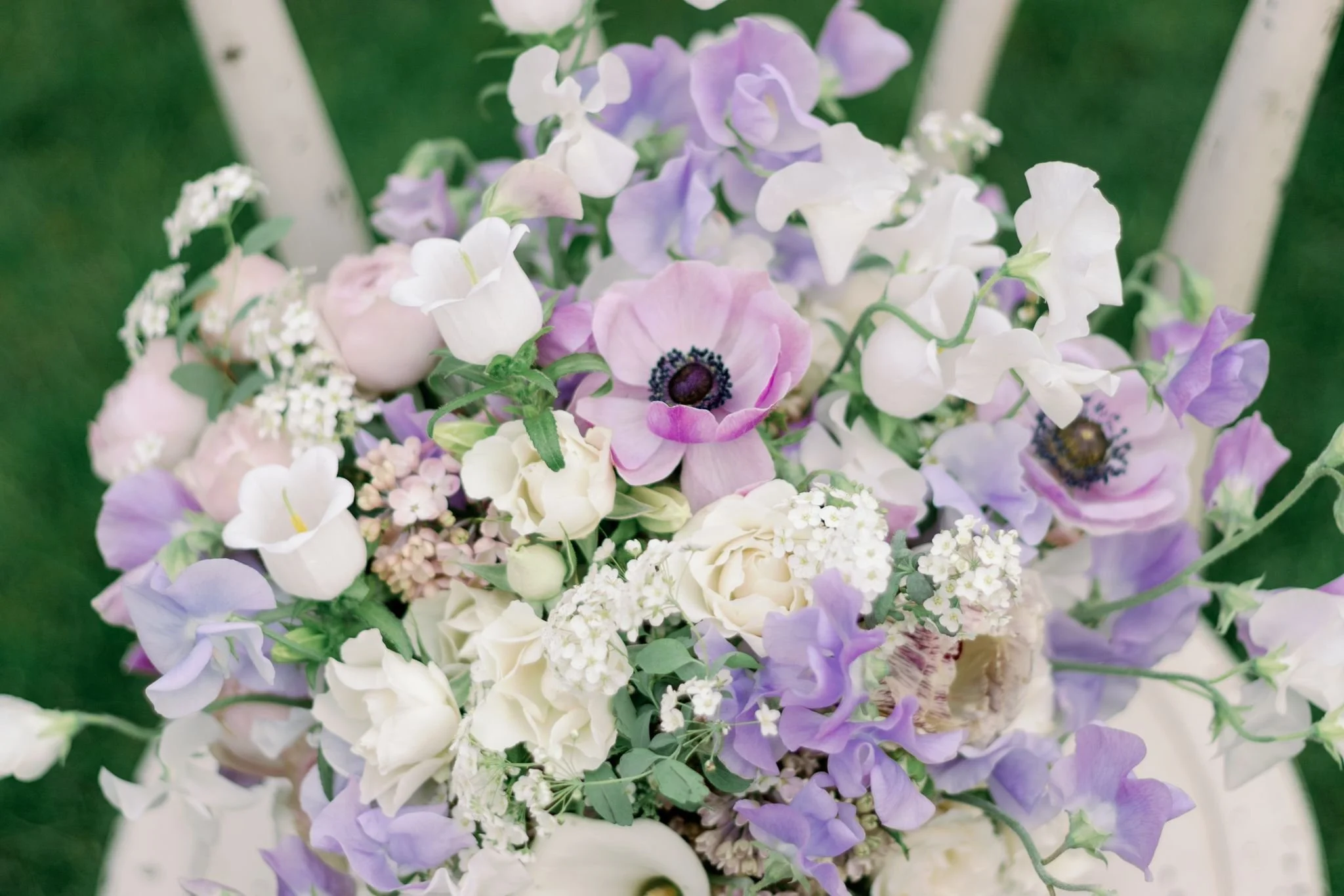 A bouquet of pastel-colored flowers, including white, pink, and purple blossoms, arranged on a white chair outdoors with greenery in the background.