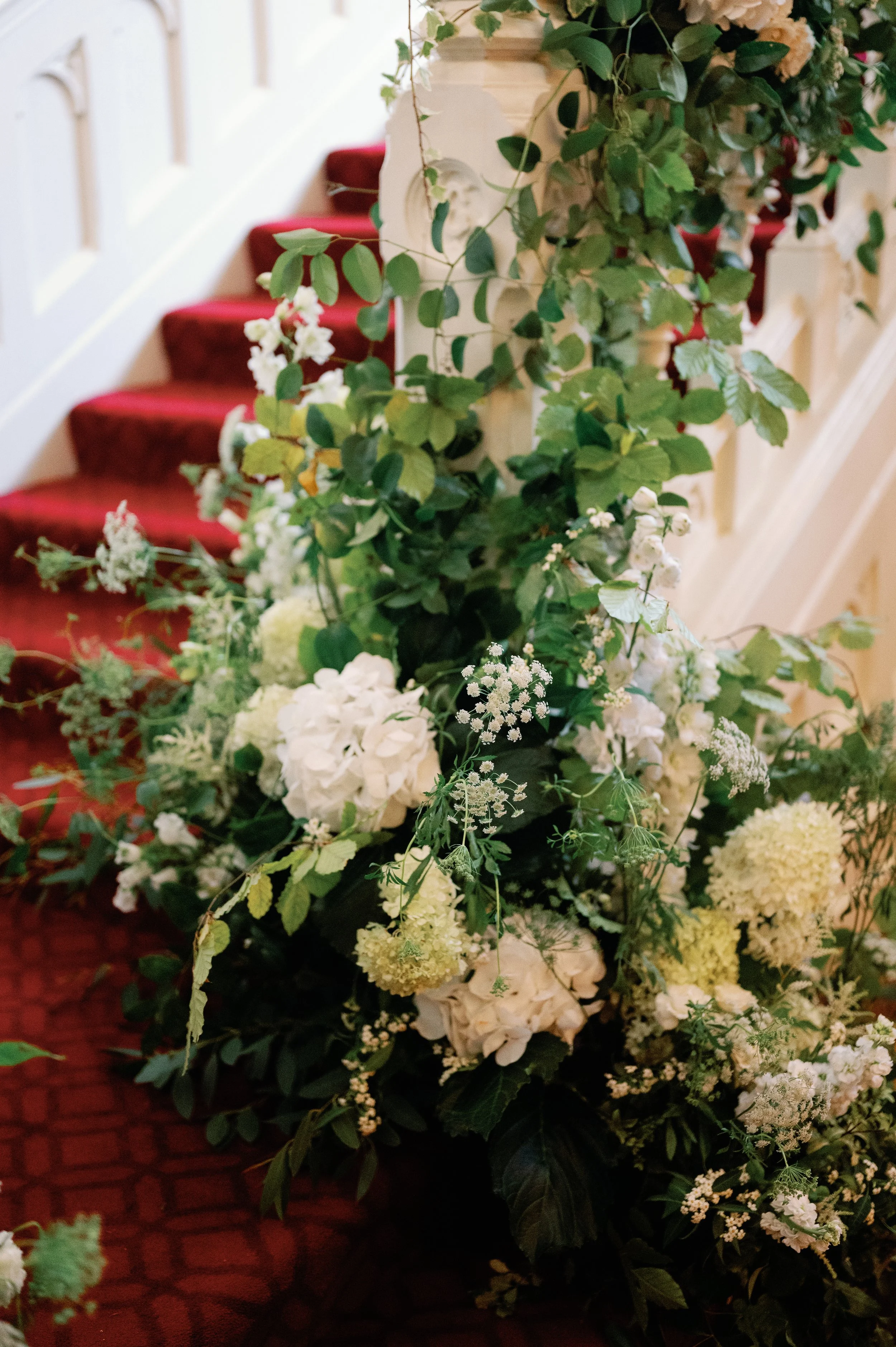 A large floral arrangement with white and green flowers and foliage, placed at the bottom of a staircase with red carpet.