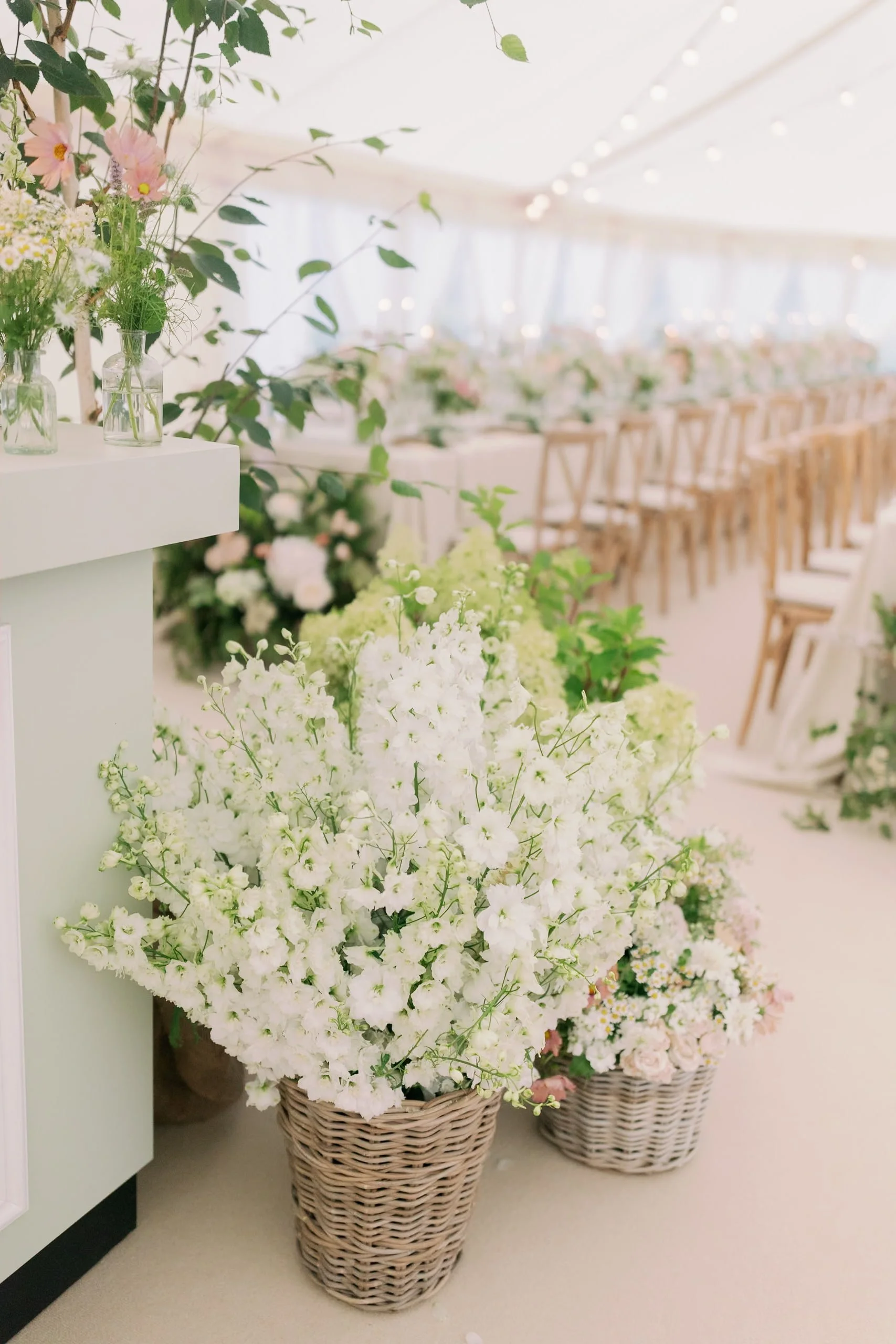 Marquee wedding reception with white floral arrangements in wicker baskets, a long dining table with pink and white flowers, wooden chairs, and fairy lights overhead. Norfolk village wedding by The Botany House Wedding Florist. Luxury wedding florals