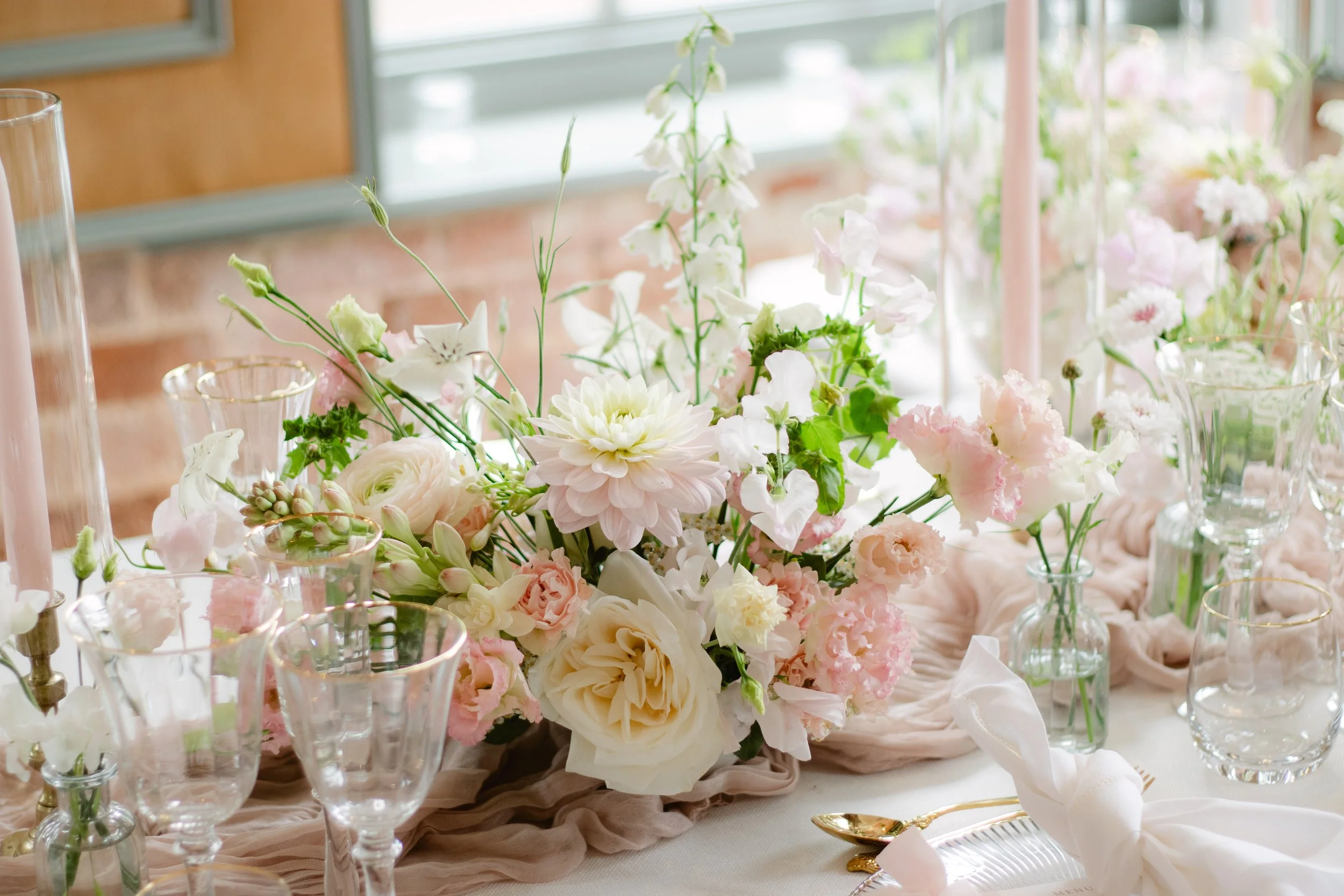 Elegant table centerpiece with pastel pink and white flowers, including roses, dahlias, and lisianthus, surrounded by glassware and gold-accented cutlery.