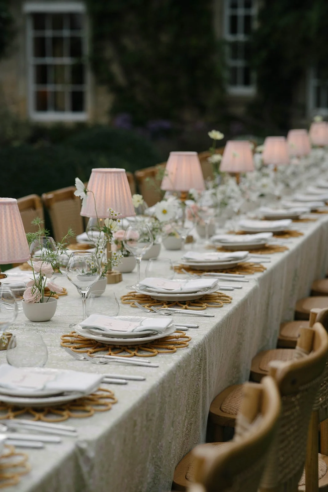 Long outdoor dining table set for a formal dinner with pink lamps, floral centerpieces and woven placemats, with wooden chairs, in a garden setting at dusk - luxury garden-style wedding flowers by The Botany House, wedding florist for Cornwell Manor