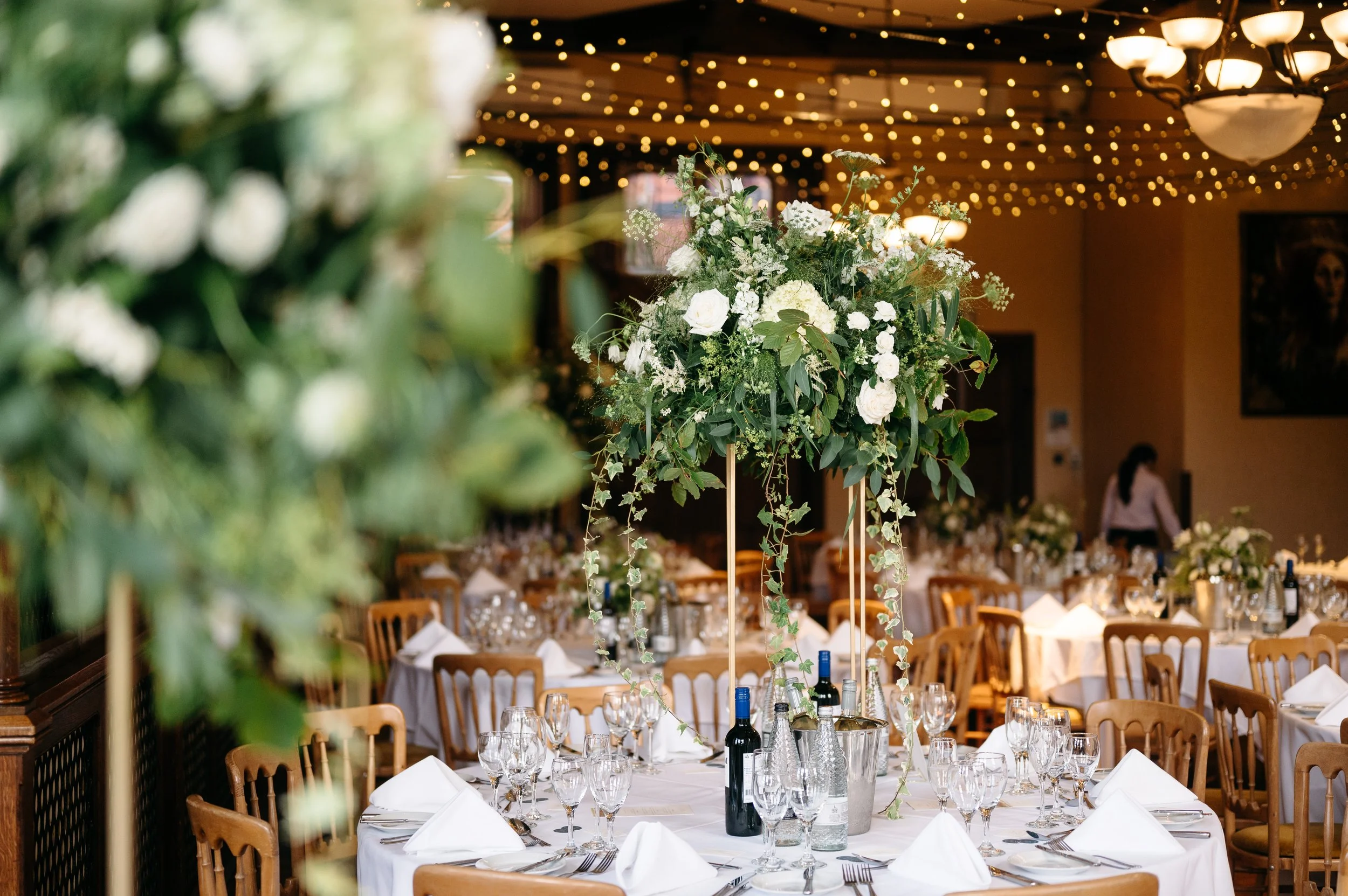 Elegant banquet table with white tablecloths, napkins, glasses, and a tall floral centerpiece with white flowers and greenery at a wedding or event venue decorated with string lights and chandeliers.
