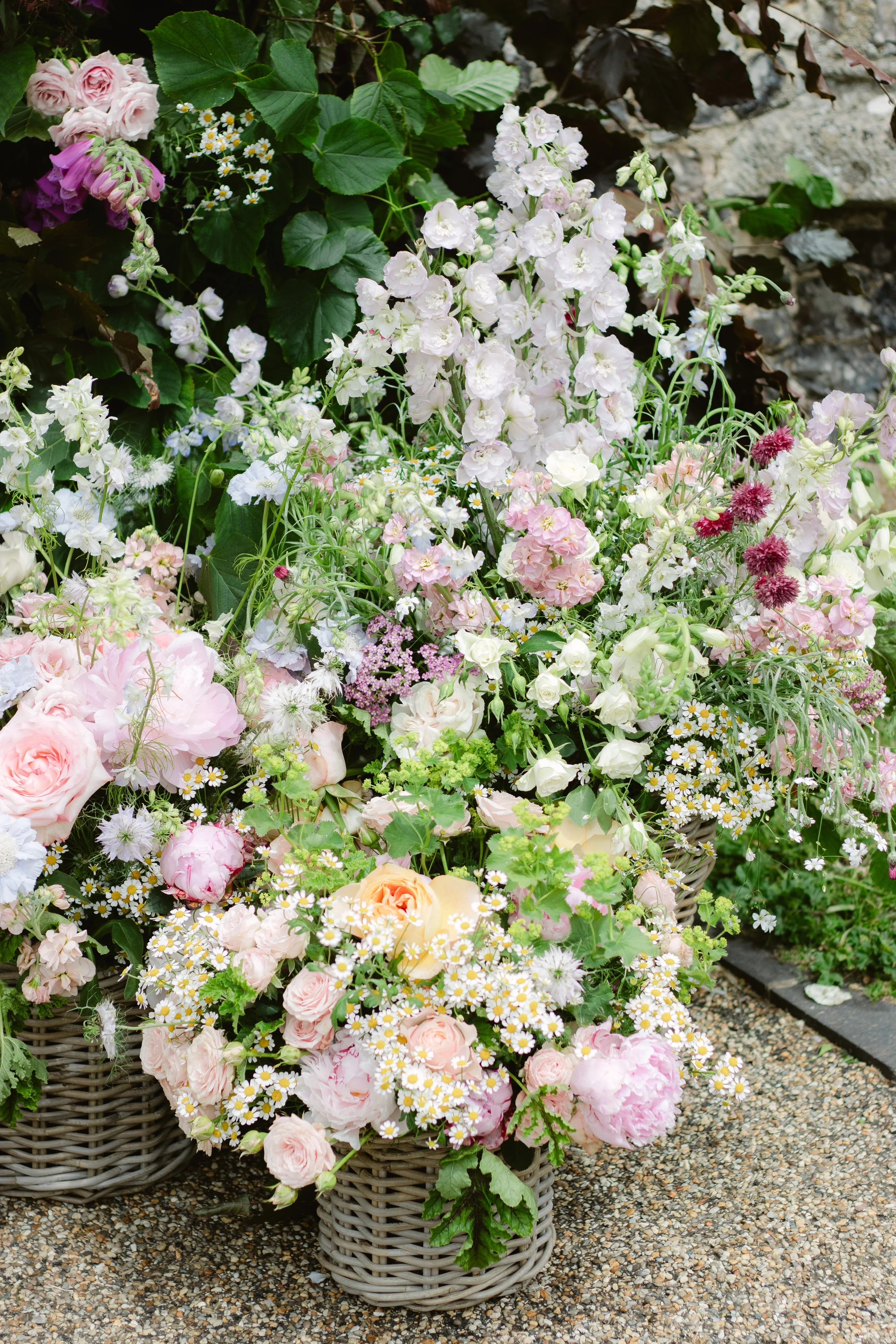 A basket of mixed pastel-colored flowers, including roses and daisies, placed on a stone surface, with a backdrop of green leaves and rocks.