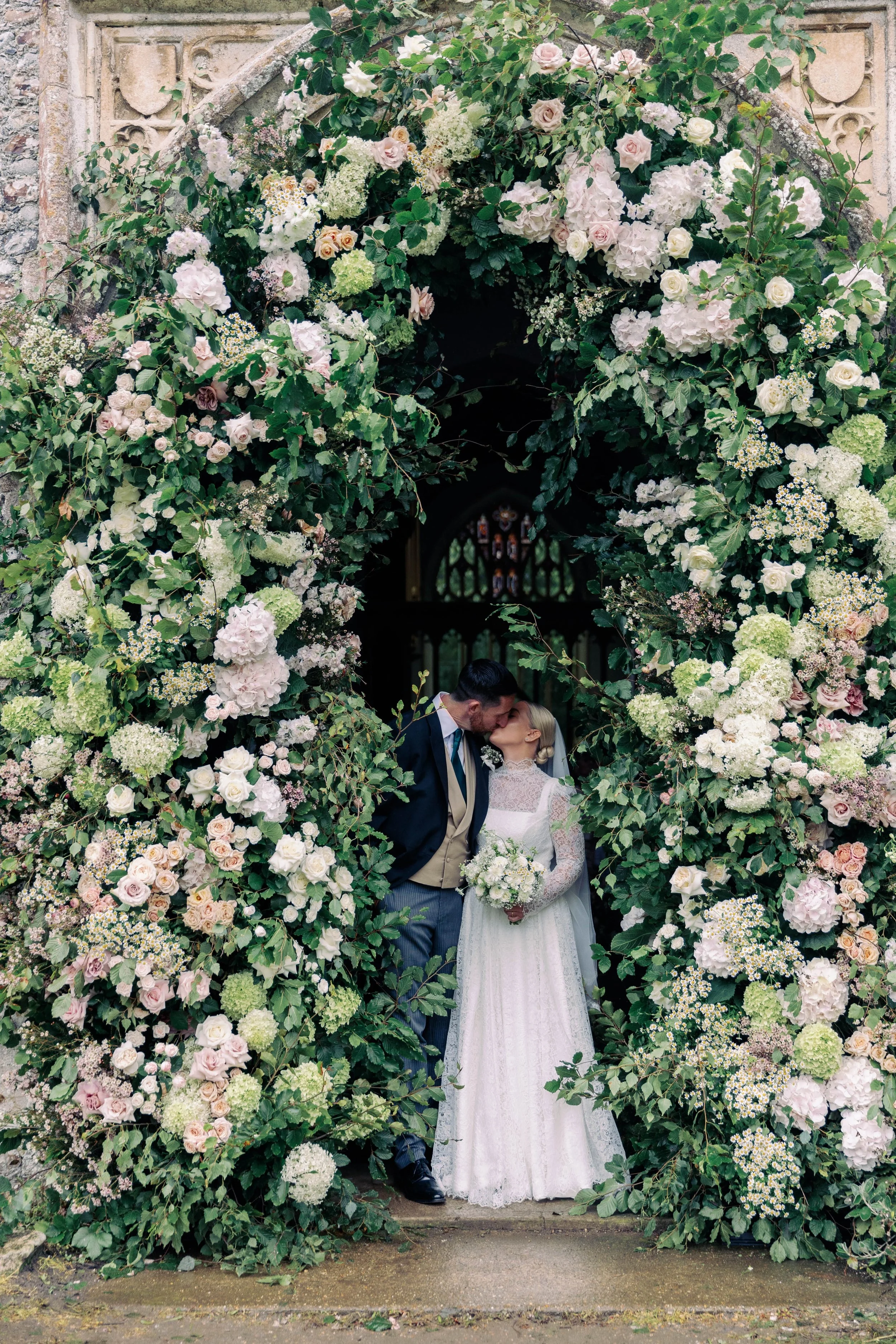 A bride and groom sharing a kiss under a large floral arch at their wedding. The arch is made of warm, pastel coloured flowers with green foliage. The bride is wearing a white lace wedding dress and holding a bouquet, while the groom is dressed in a