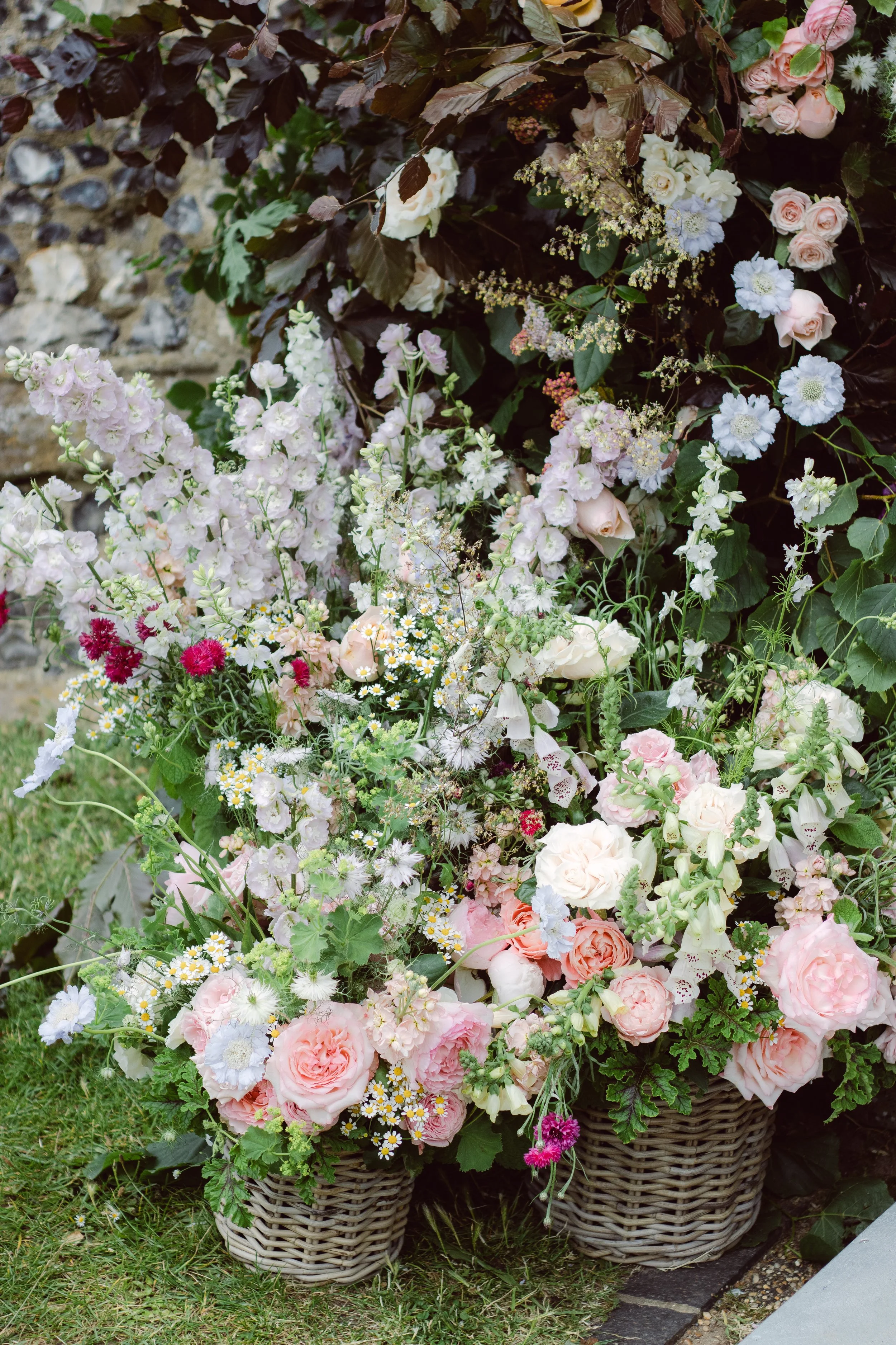Two wicker baskets filled with pink, white, and red flowers, including roses and various small blooms, placed outdoors near a stone wall and green grass.