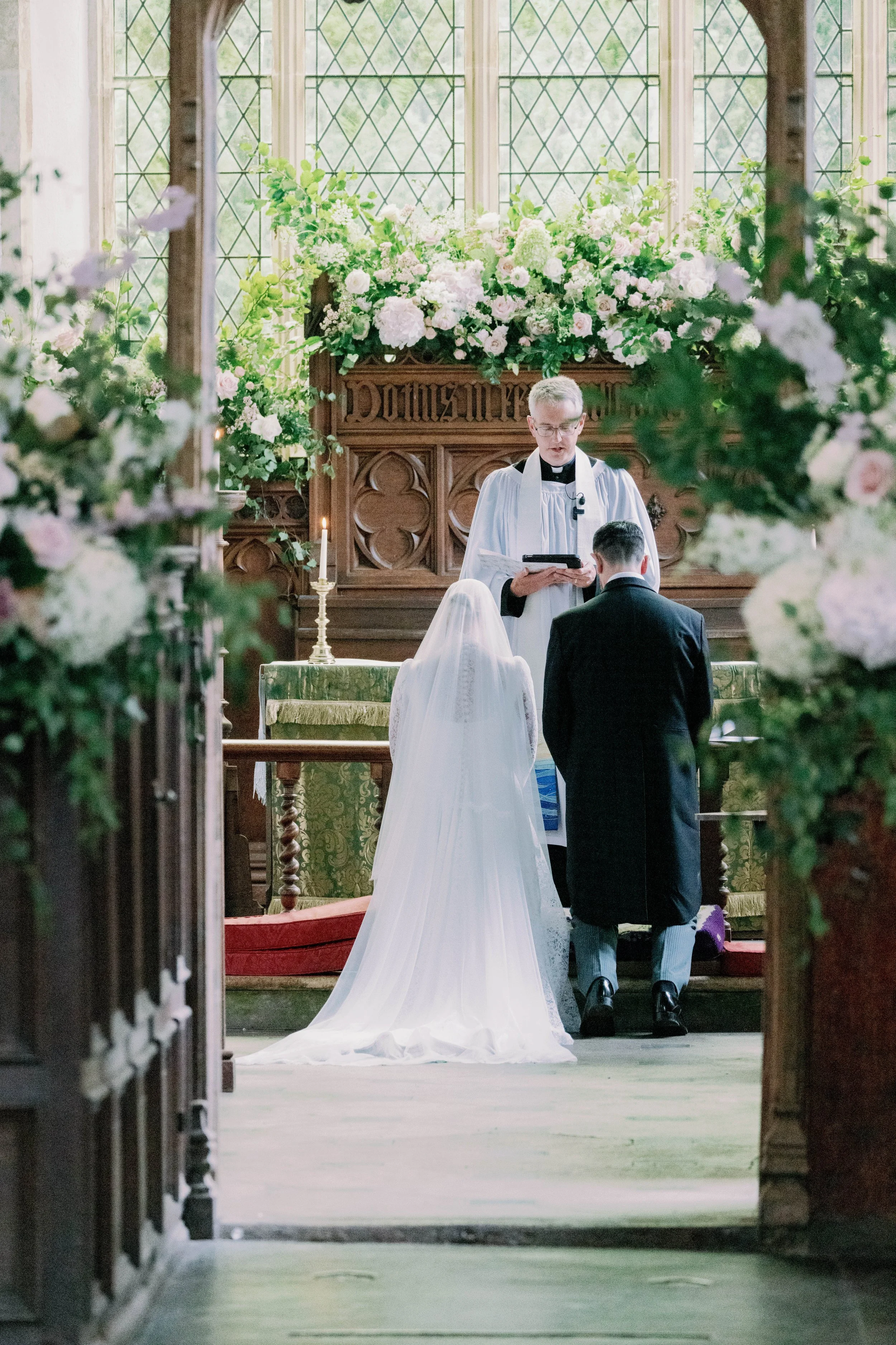 A wedding ceremony taking place in a church with a priest officiating, a bride in a white veil kneeling, and a groom in a black suit kneeling before the altar, surrounded by garden style floral arrangements