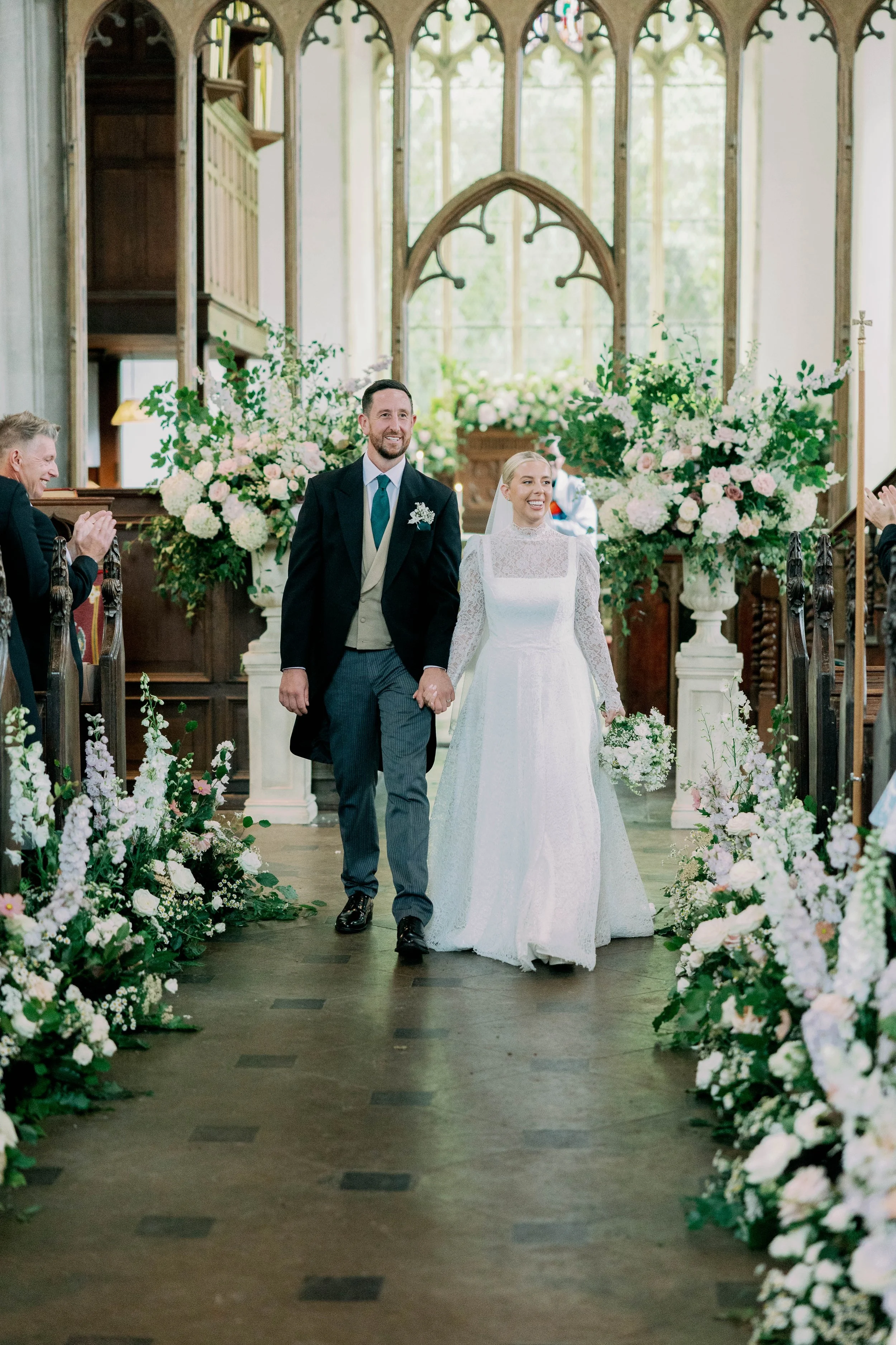 Bride and groom walking down the aisle at a wedding ceremony inside a historic church with stained glass windows and floral arrangements.