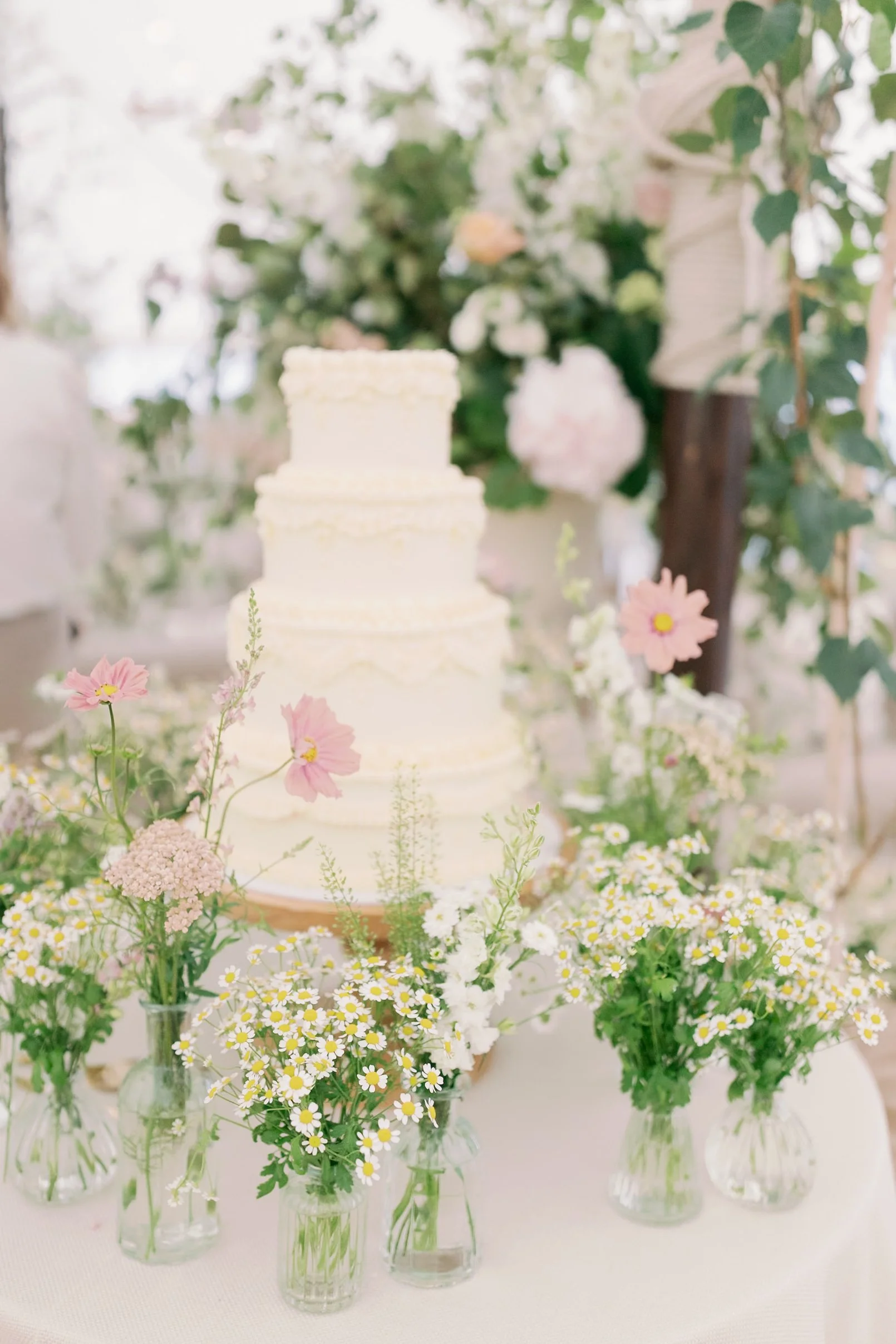 A three-tier white wedding cake surrounded by small glass vases filled with garden style flowers, set on a table with a pink cream, with a floral backdrop.