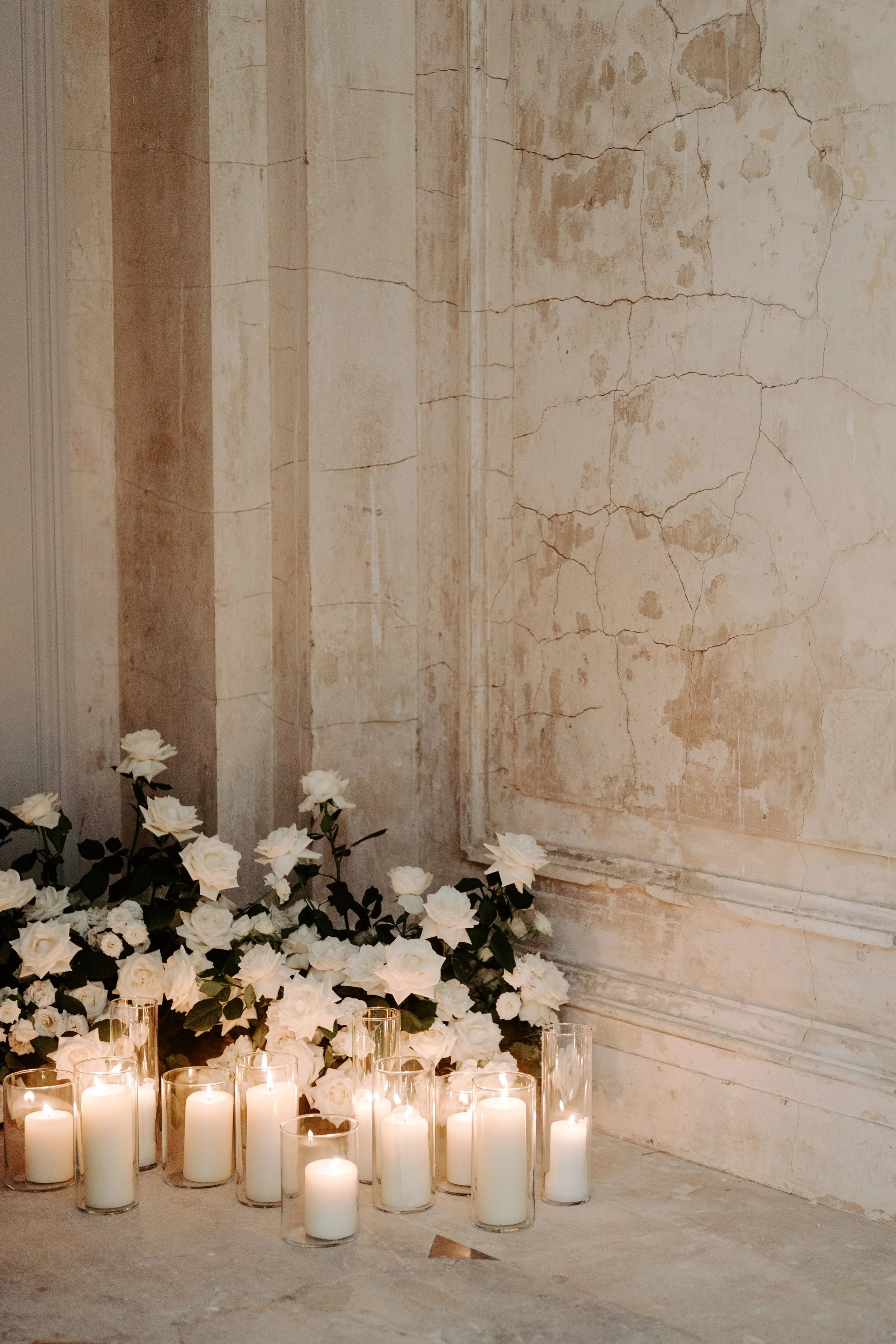 A collection of white candles in glass holders arranged on the floor, surrounded by white roses and greenery, against a rustic, peeling wall.