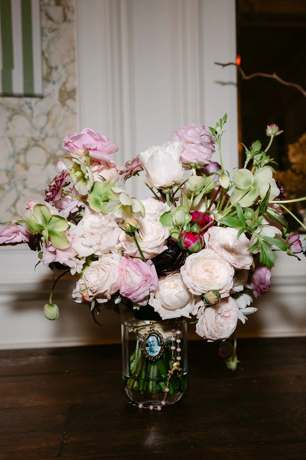 A bouquet of pink, white, and purple flowers in a glass vase on a dark wooden table, with a small religious pendant hanging on the vase.