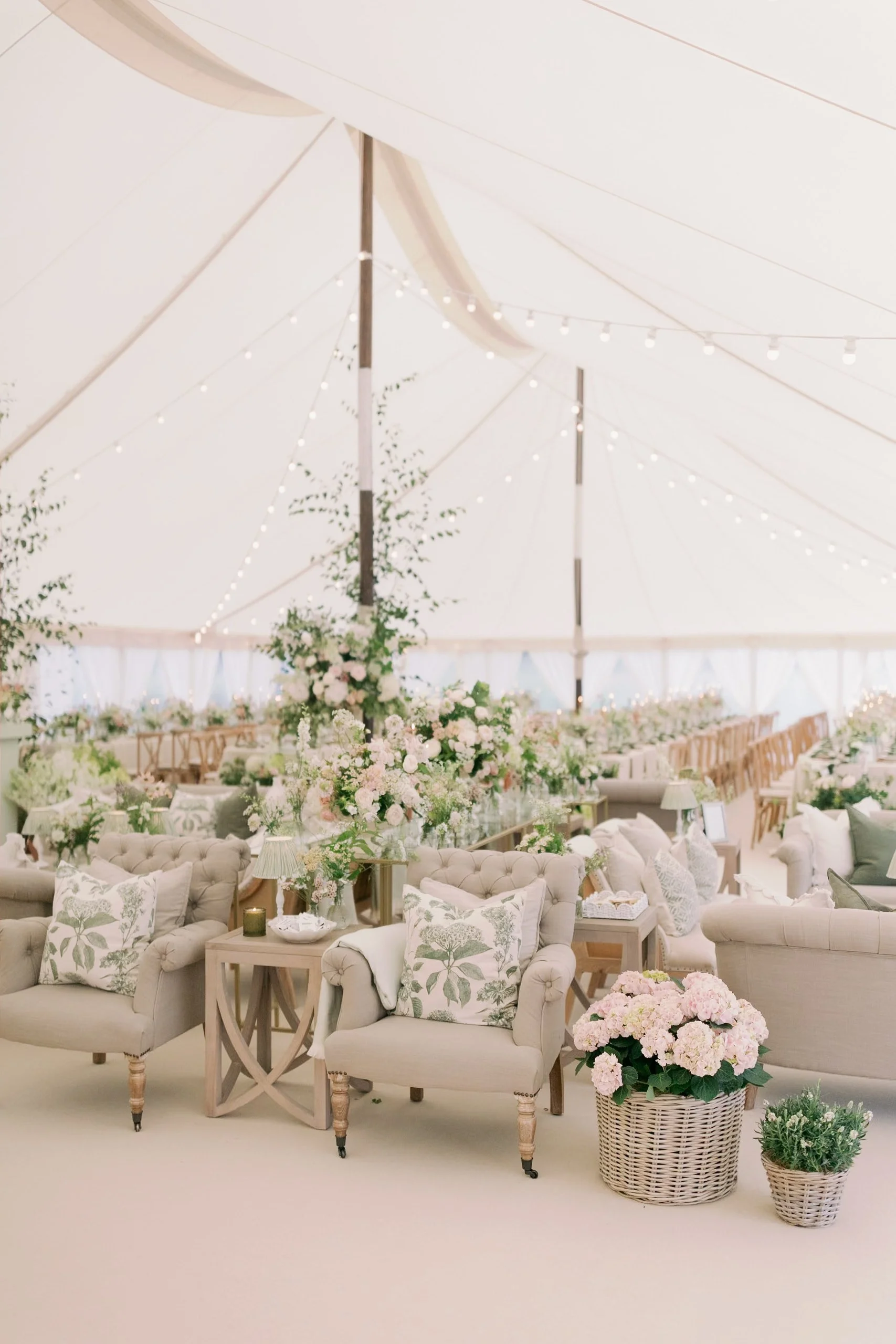 Elegant wedding reception inside a large tent with floral arrangements, cozy armchairs, and string lights. Norfolk village wedding by The Botany House Wedding Florist. Luxury wedding florals