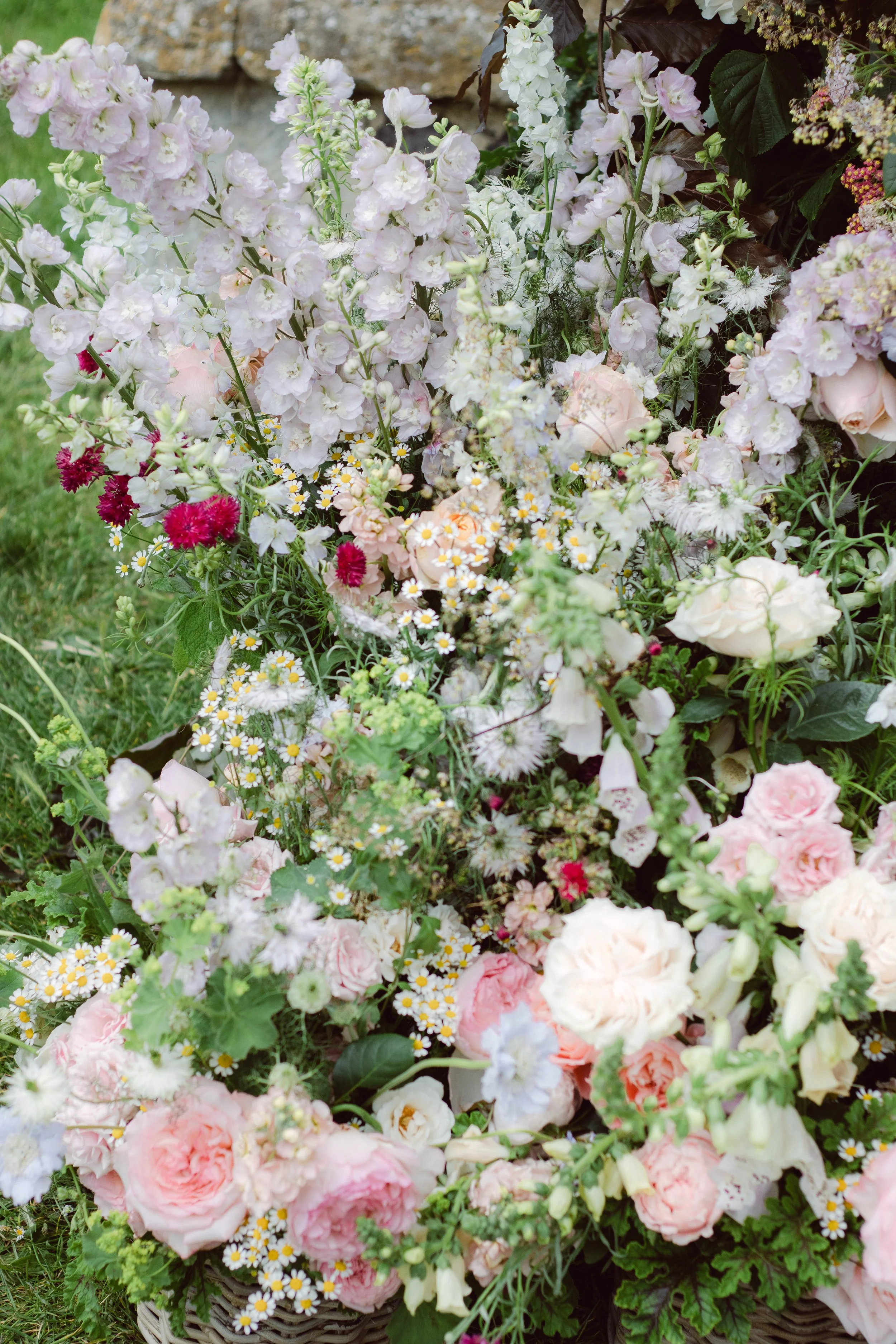 Arrangement of pink and white flowers, including roses, daisies, and delphiniums, with some greenery and dark leaves.