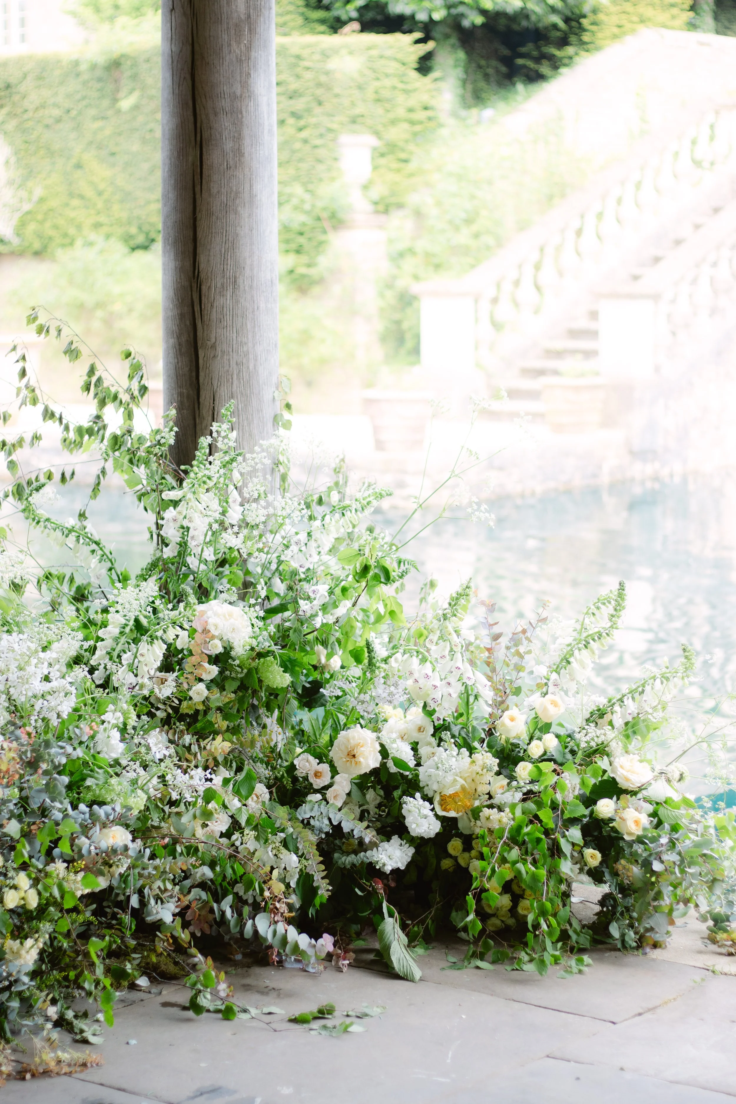 A floral arrangement of white and green flowers and foliage next to a wooden post near a body of water, with a stone staircase and greenery in the background.