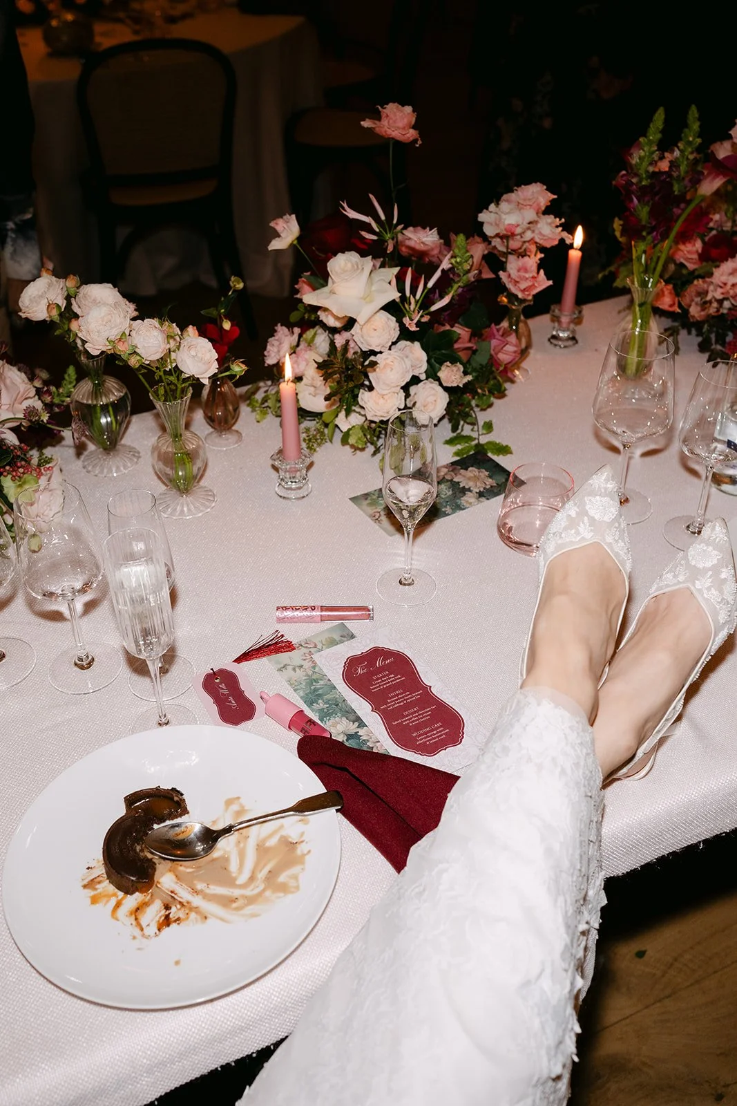 Close-up of a decorated wedding reception table with flowers, candles, wine glasses, and a plate with leftover dessert, a person's feet in lace shoes resting on the edge of the table.