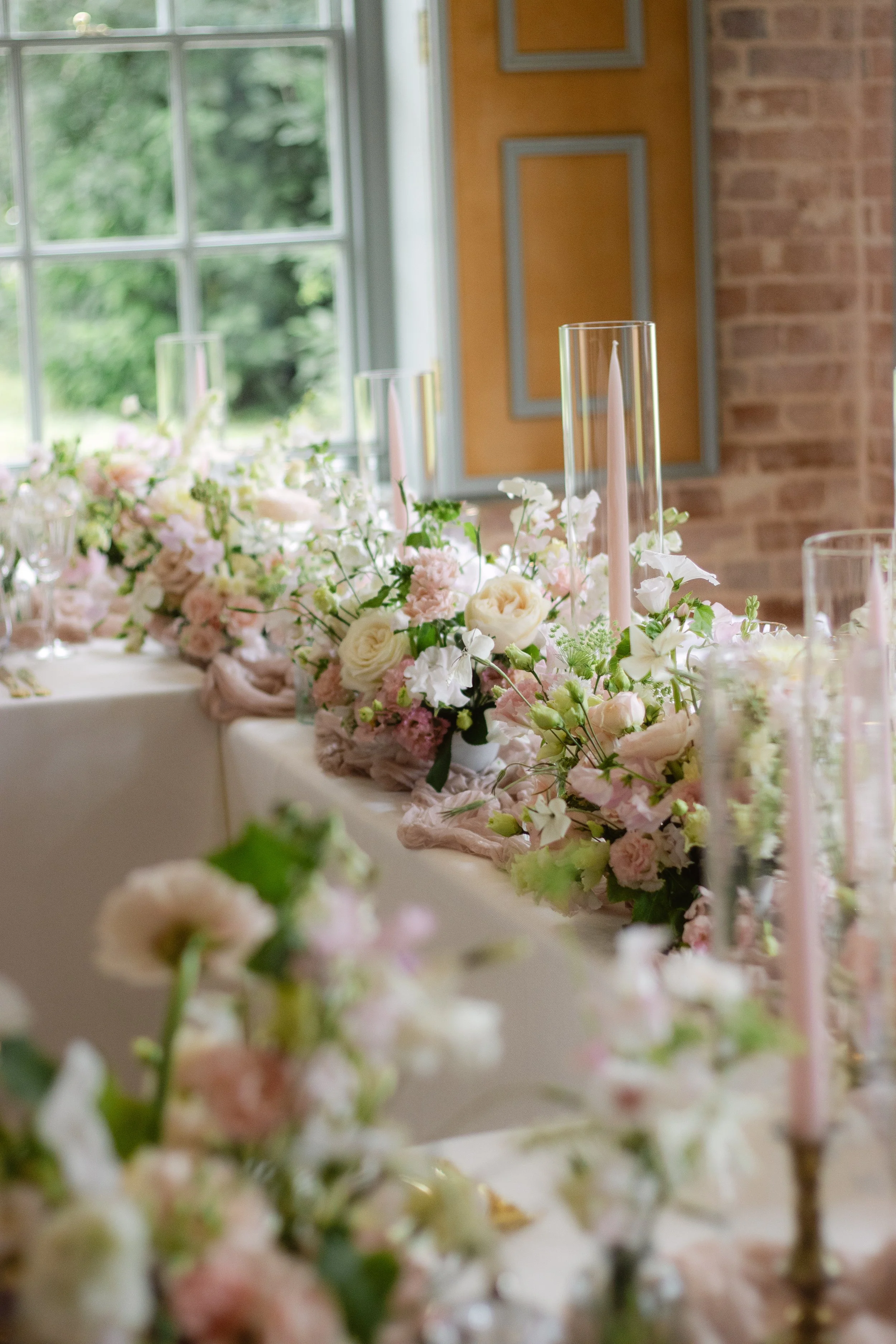 Indoor wedding or event table decorated with pastel pink and white flowers, tall glass candle holders, and pink candles, in front of a window with greenery outside and a brick wall.