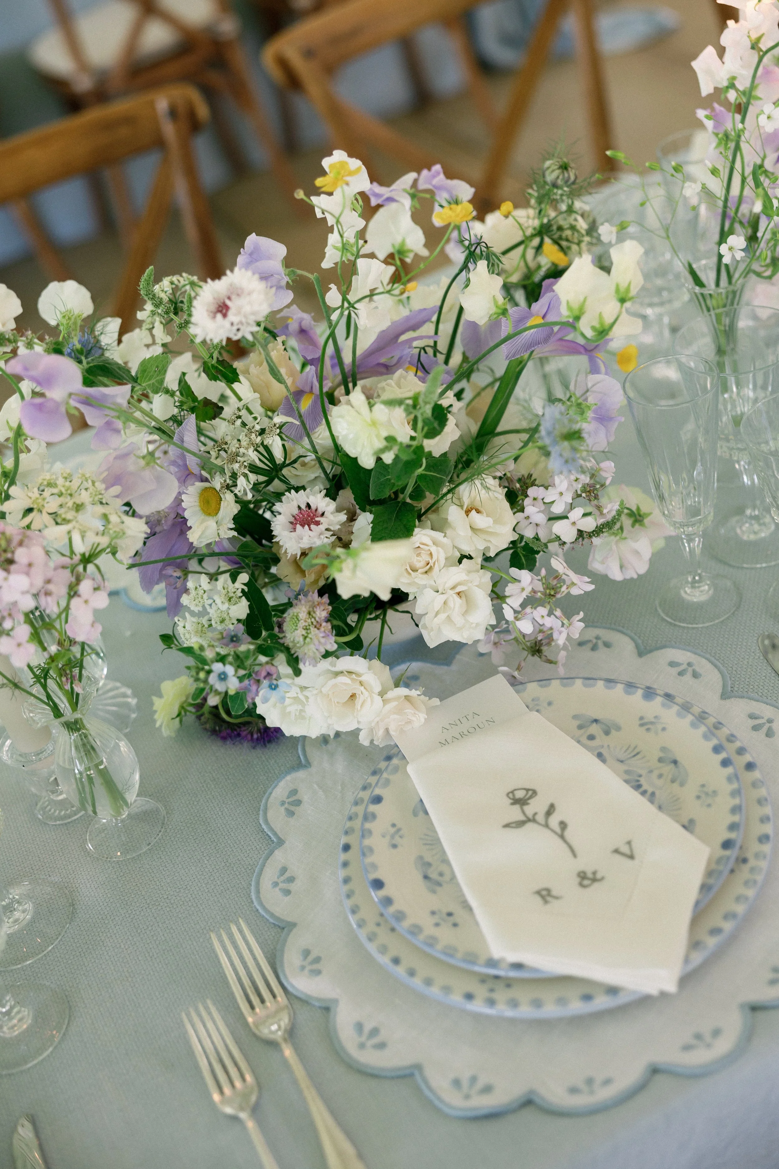 Elegant table setting with a pastel-colored floral centerpiece, patterned blue and white plates, clear glassware, and silver forks, at a formal event or wedding reception.