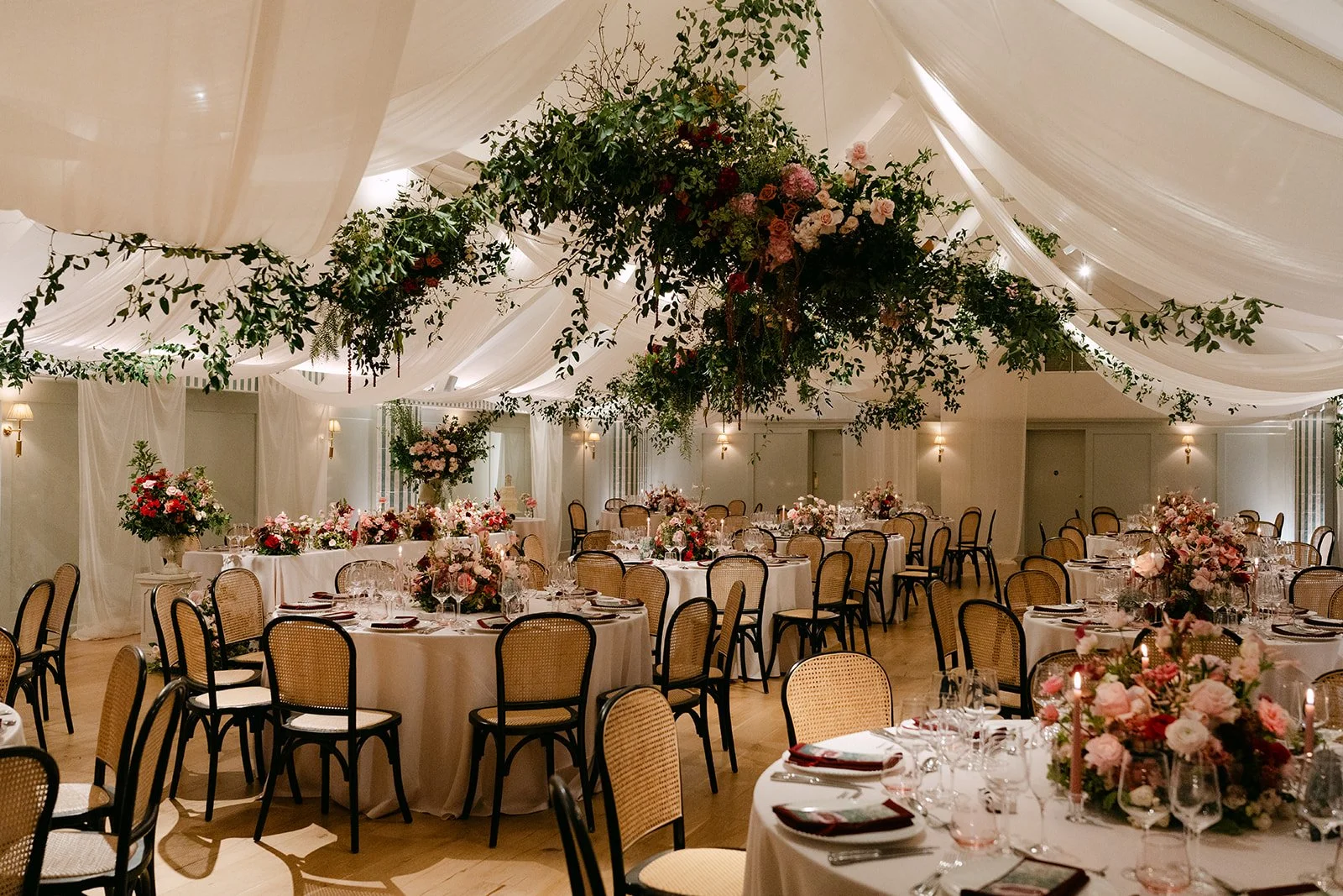 An elegant banquet hall decorated for a wedding or special event with round tables covered with white tablecloths, pink and white floral centerpieces, and black chairs. The ceiling features hanging white drapes and lush floral arrangements with green