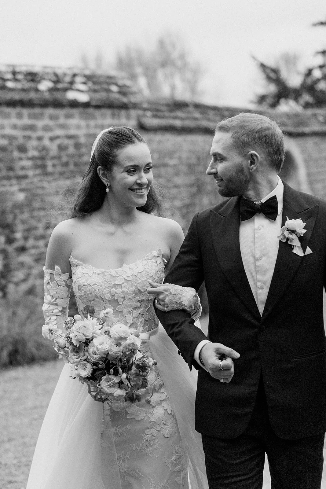 A bride and groom walking arm in arm outdoors, smiling at each other, with the bride holding a bouquet of flowers and wearing a lace wedding dress, and the groom in a tuxedo with a boutonniere, in a black-and-white photo. The Botany House Wedding