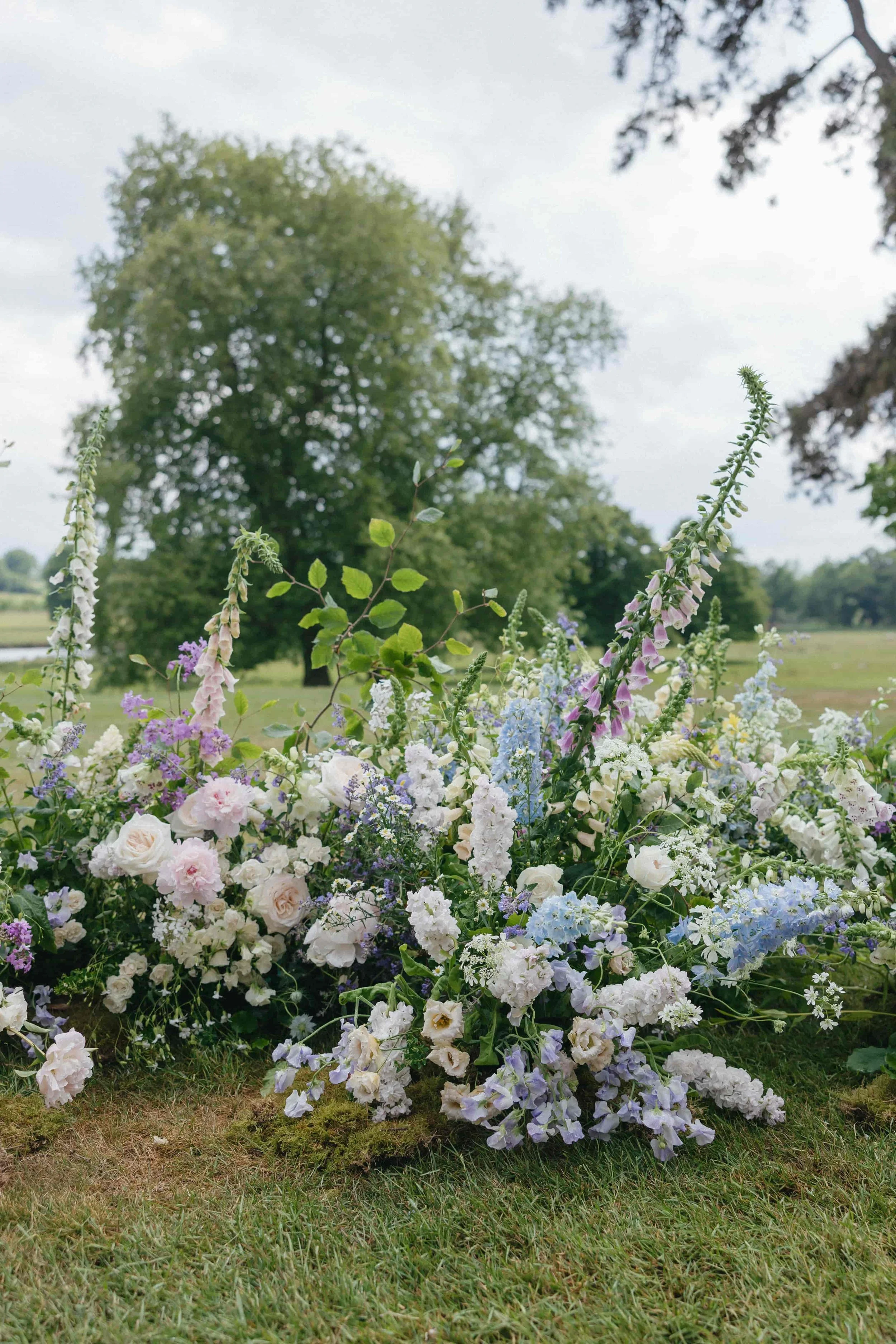 Colourful garden-inspired wedding florals with roses, delphiniums, and wildflowers on grass at Suffolk wedding. Florals By The Botany House. Planning by Attabara Studio