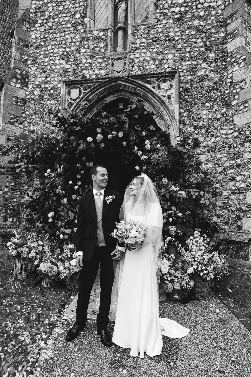 A bride in a white wedding gown and a groom in a black suit holding hands and smiling at each other outside a church decorated with flowers and greenery.