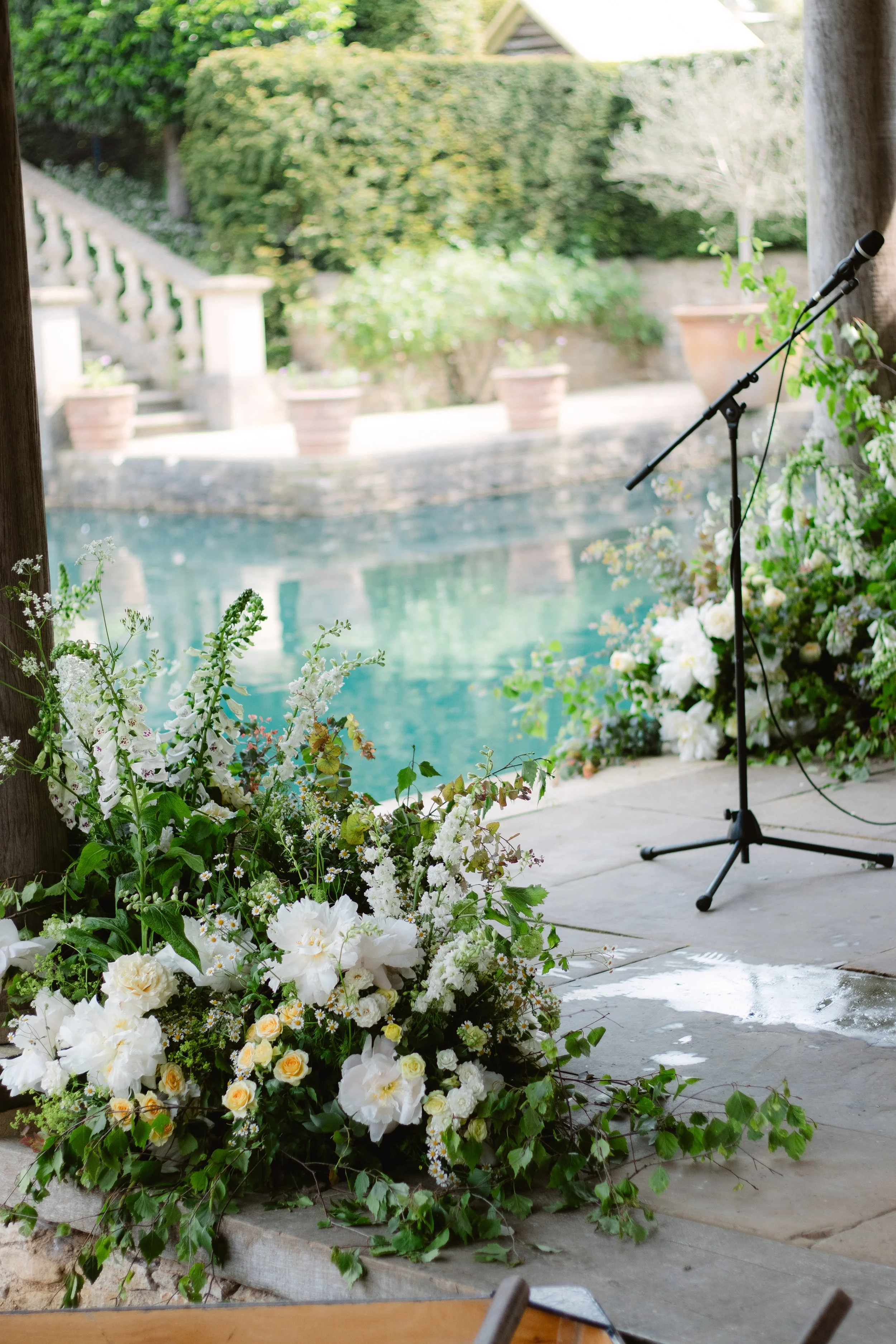 Floral arrangement with white flowers and greenery on a stage setting near a pool, with a microphone stand, trees, and potted plants in the background.