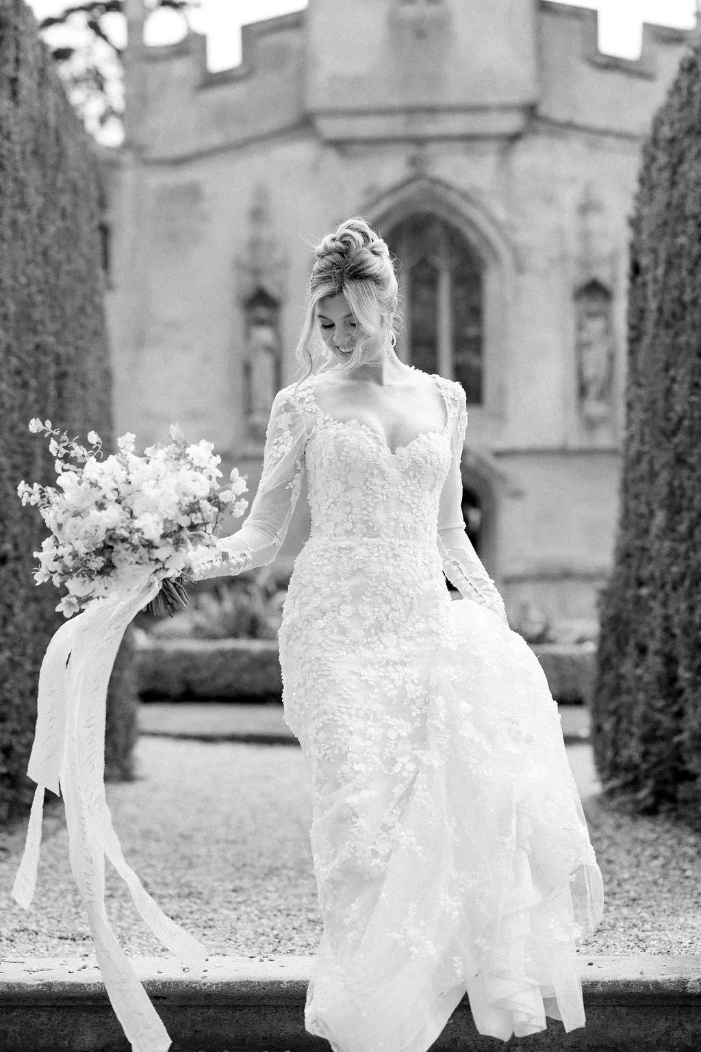 Black and white photo of a woman in a wedding dress holding a bouquet of flowers outdoors in front of a historic building.