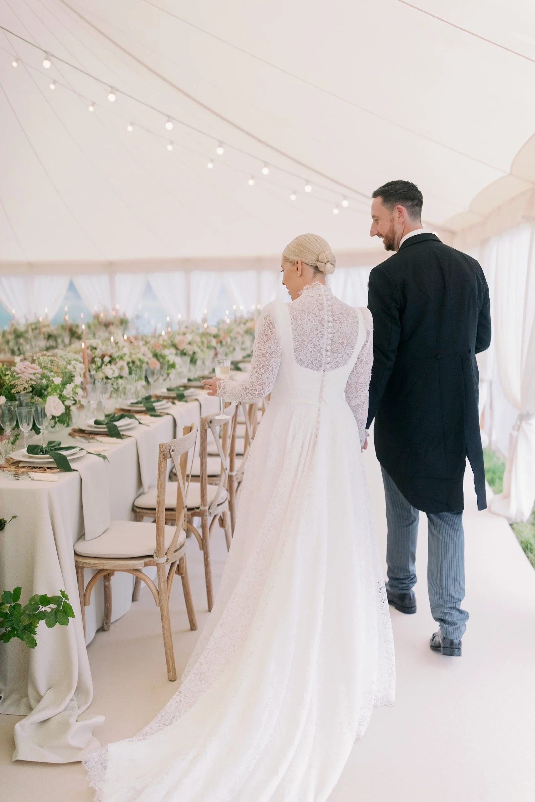 A bride and groom walking away from the camera at a decorated wedding reception sail cloth marquee, with the bride in a white lace wedding gown and the groom in a black tailcoat and striped pants.
