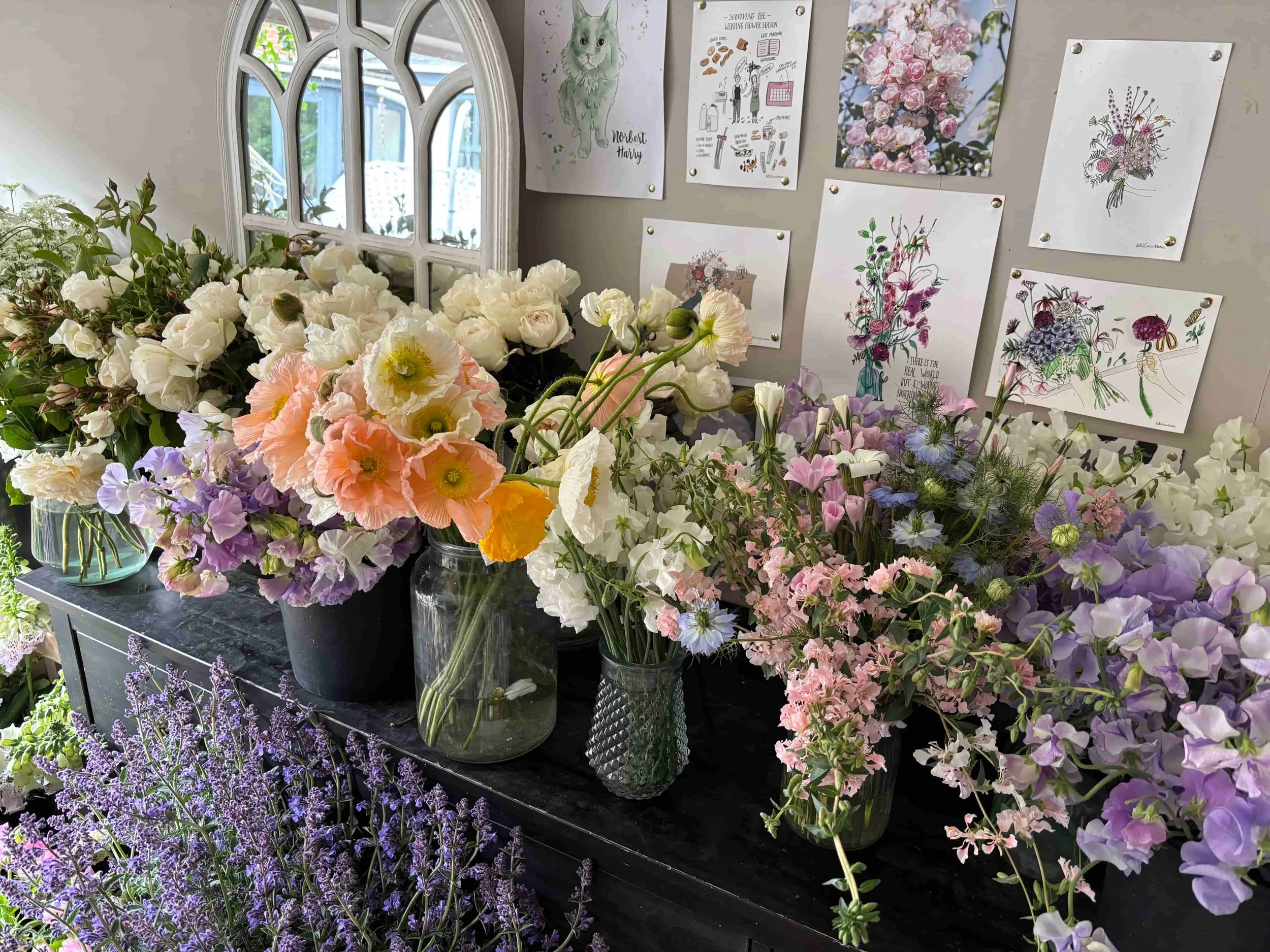 An indoor display of various fresh flowers in glass jars and containers on a black table, with a decorated window and framed floral and botanical artwork on the wall in the background. The Botany House studio in Norfolk. 