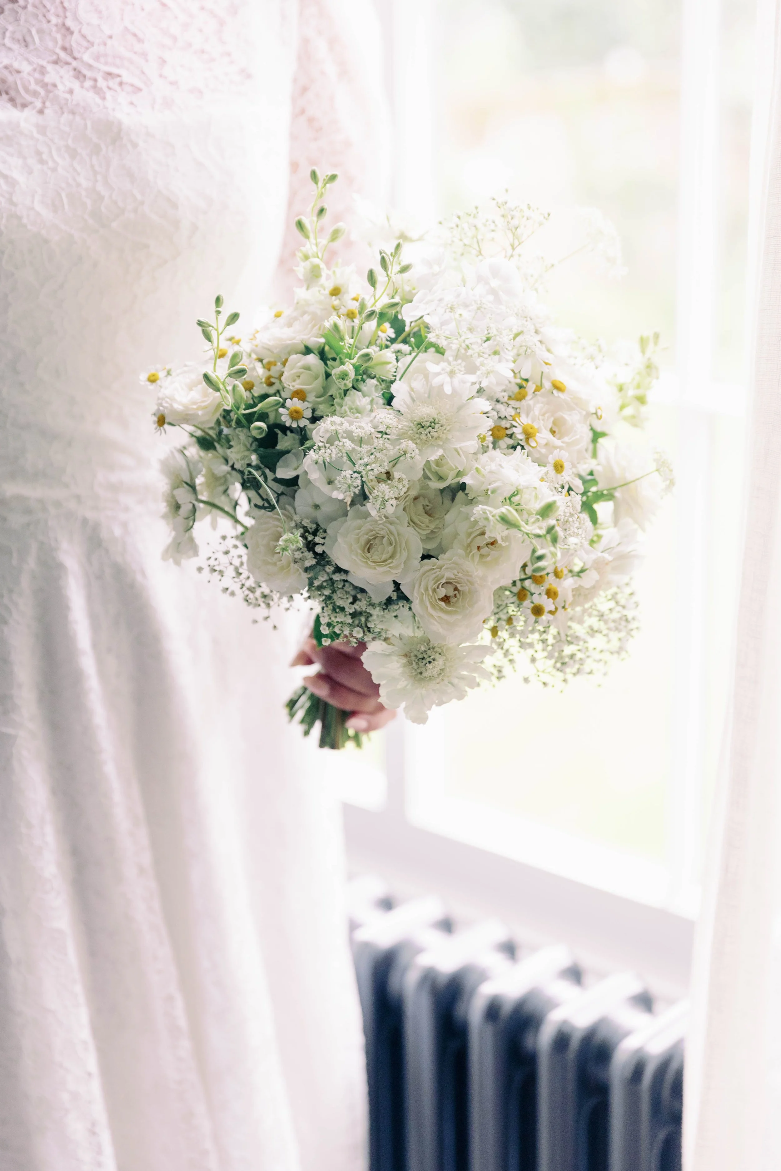 Bride holding a bouquet of white flowers near a window with a radiator below. White lace custom Emma Beaumont Dress