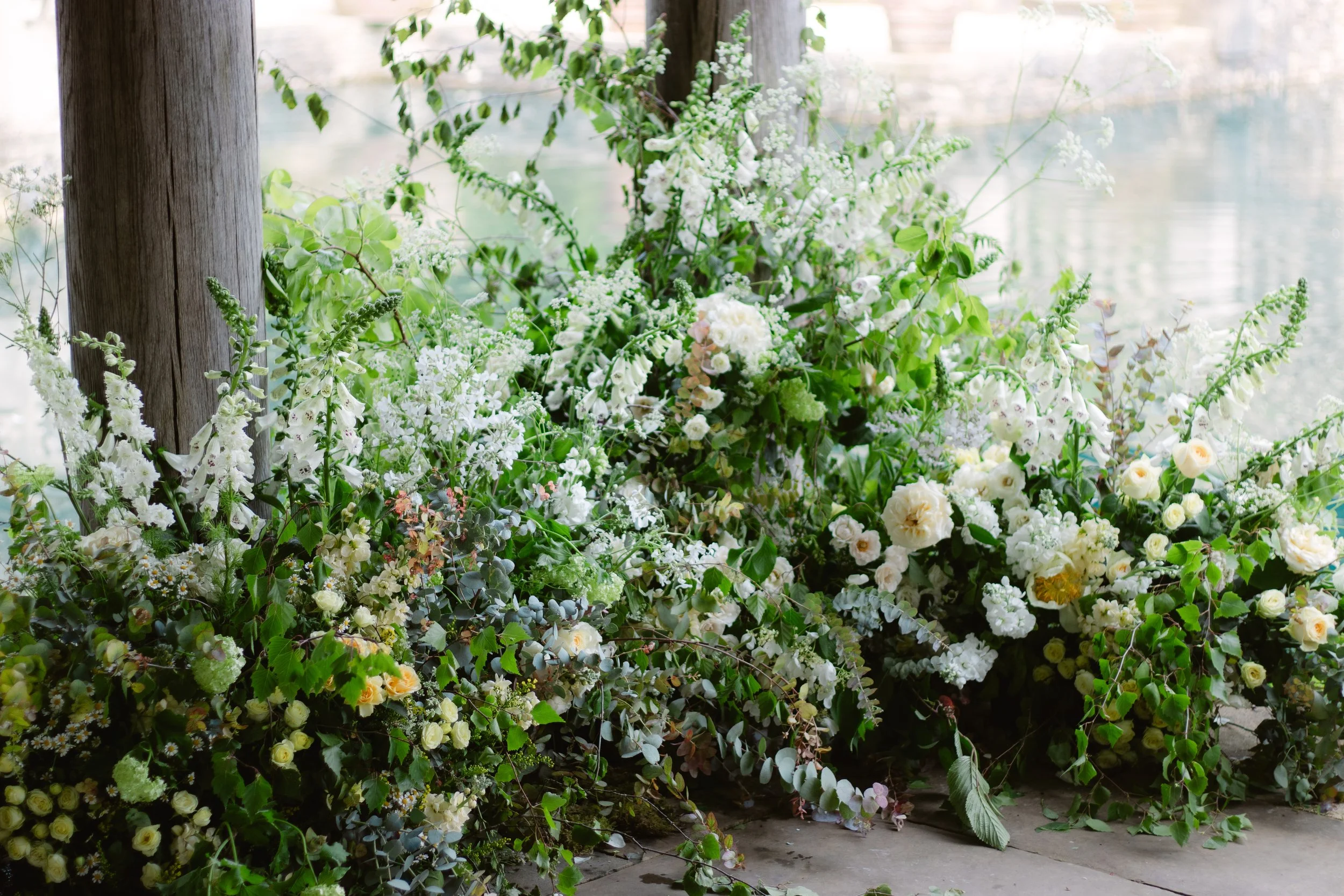 A large arrangement of white and green flowers and foliage leaning against wooden posts near a body of water.