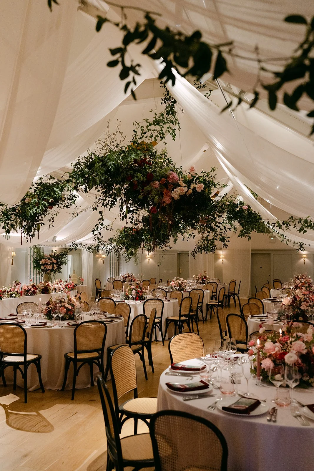 Elegant banquet hall decorated with hanging floral arrangements and draped fabric ceiling, set up for a wedding reception with round tables, floral centerpieces, candles, and place settings.