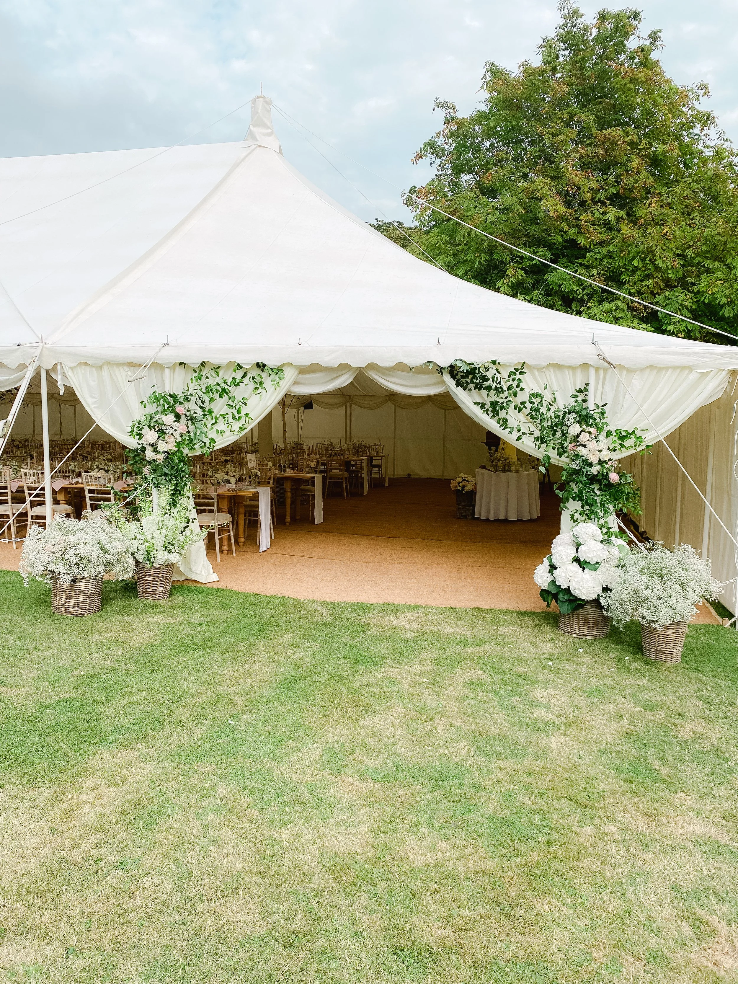 White wedding tent decorated with white and pink flowers and greenery, set up outdoors on grass with trees in the background.