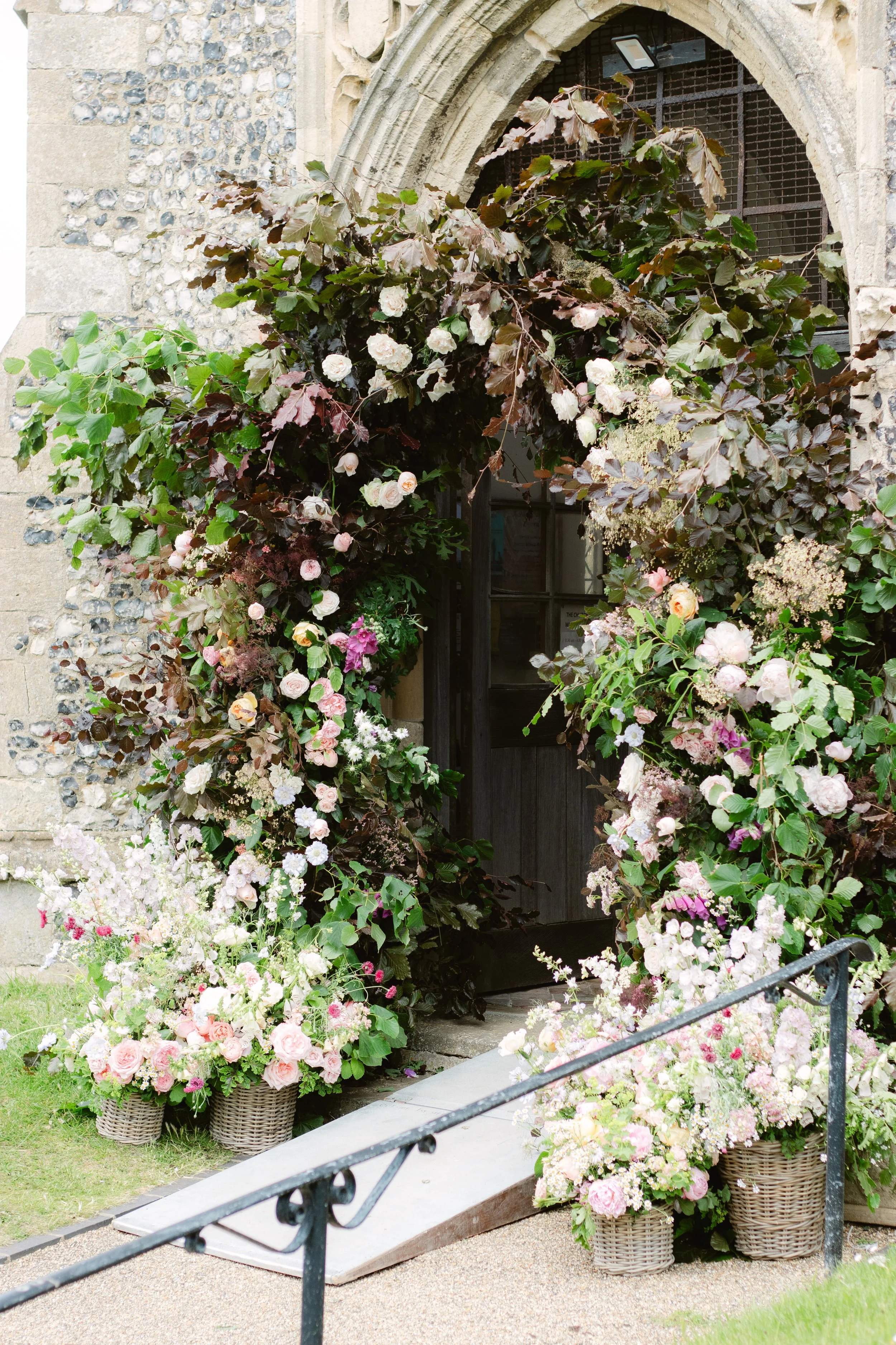 Flower arch with pink and white flowers surrounding entrance to a historic stone building, with a ramp leading to the door.
