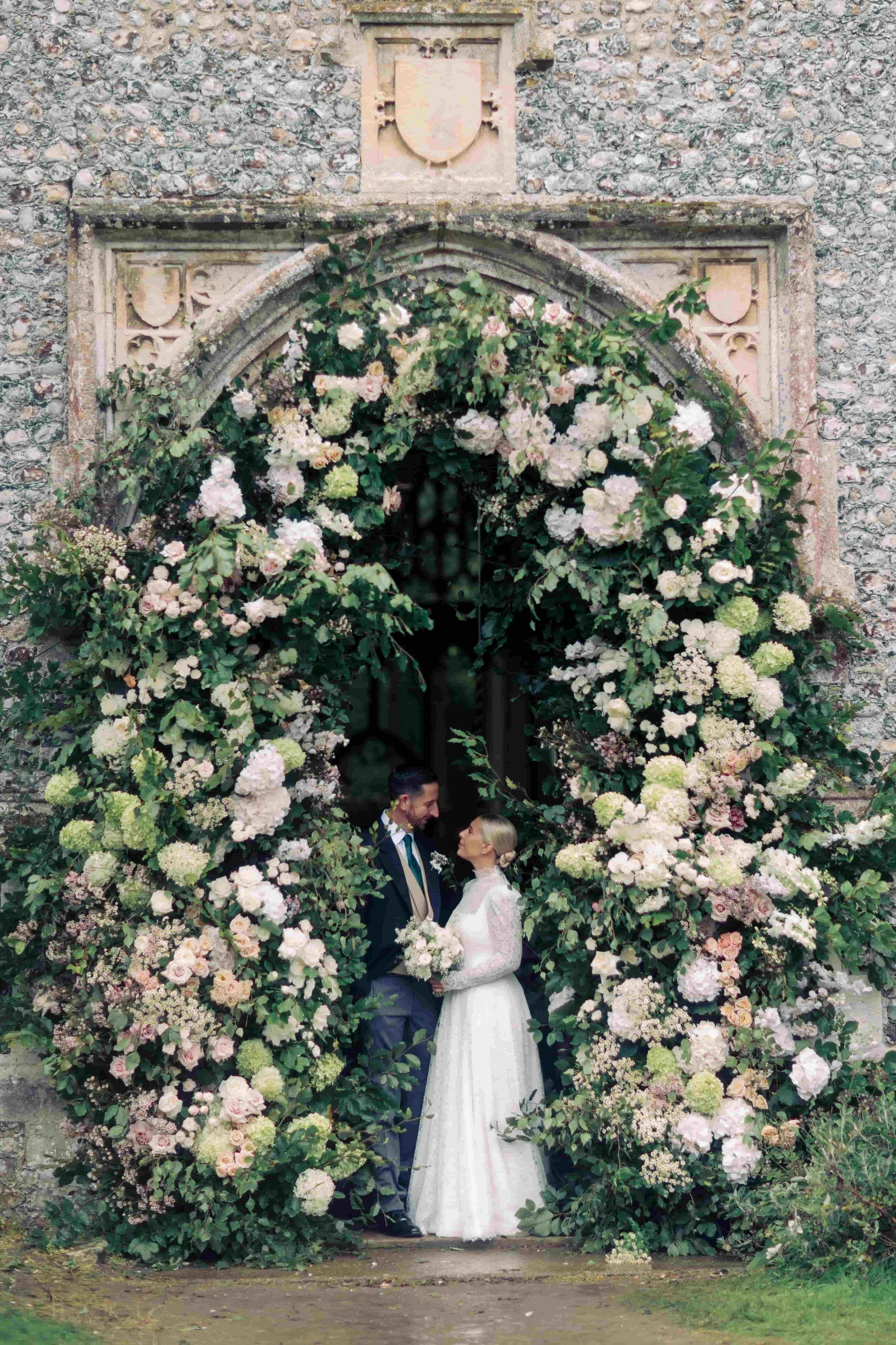 A bride and groom stand under an archway decorated with white and pink roses, outside a stone church. Norfolk village wedding by The Botany House Wedding Florist. Luxury wedding florals