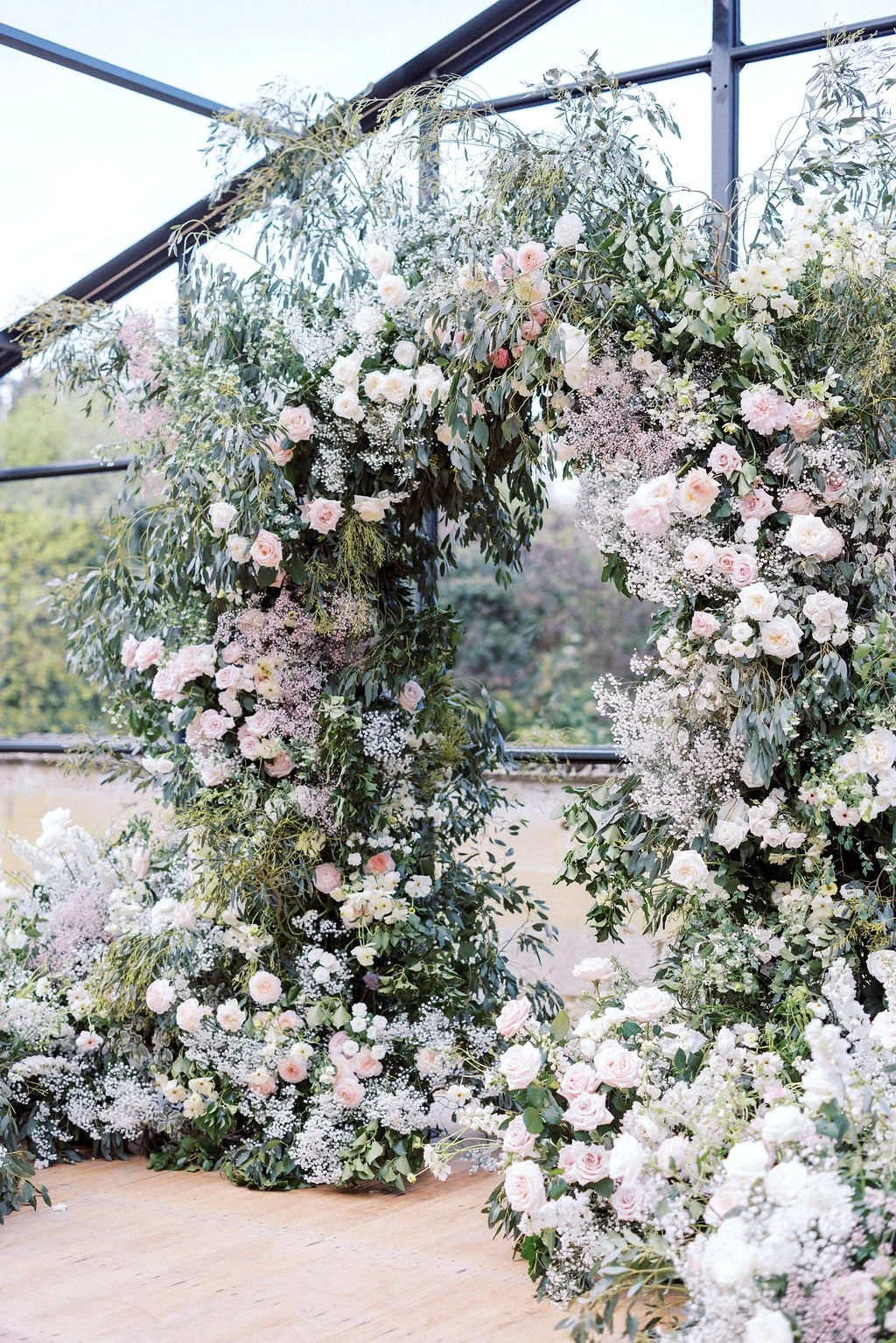 A floral arch decorated with pink and white roses, white baby's breath, and assorted greenery, set inside a greenhouse for a wedding or special event.