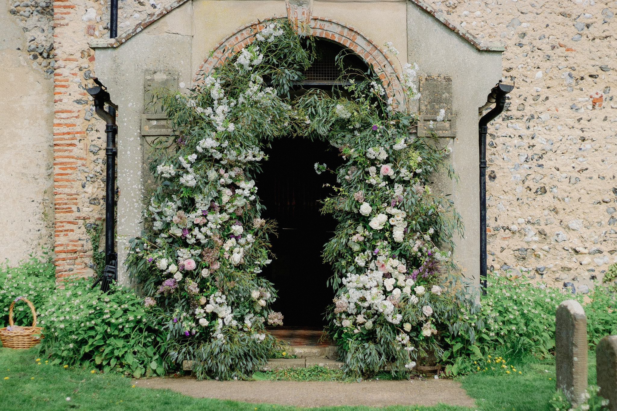 Entrance of a rustic stone and brick building decorated with a large floral arch made of white and pink flowers with green foliage, and a small stone pathway leading up to it.
